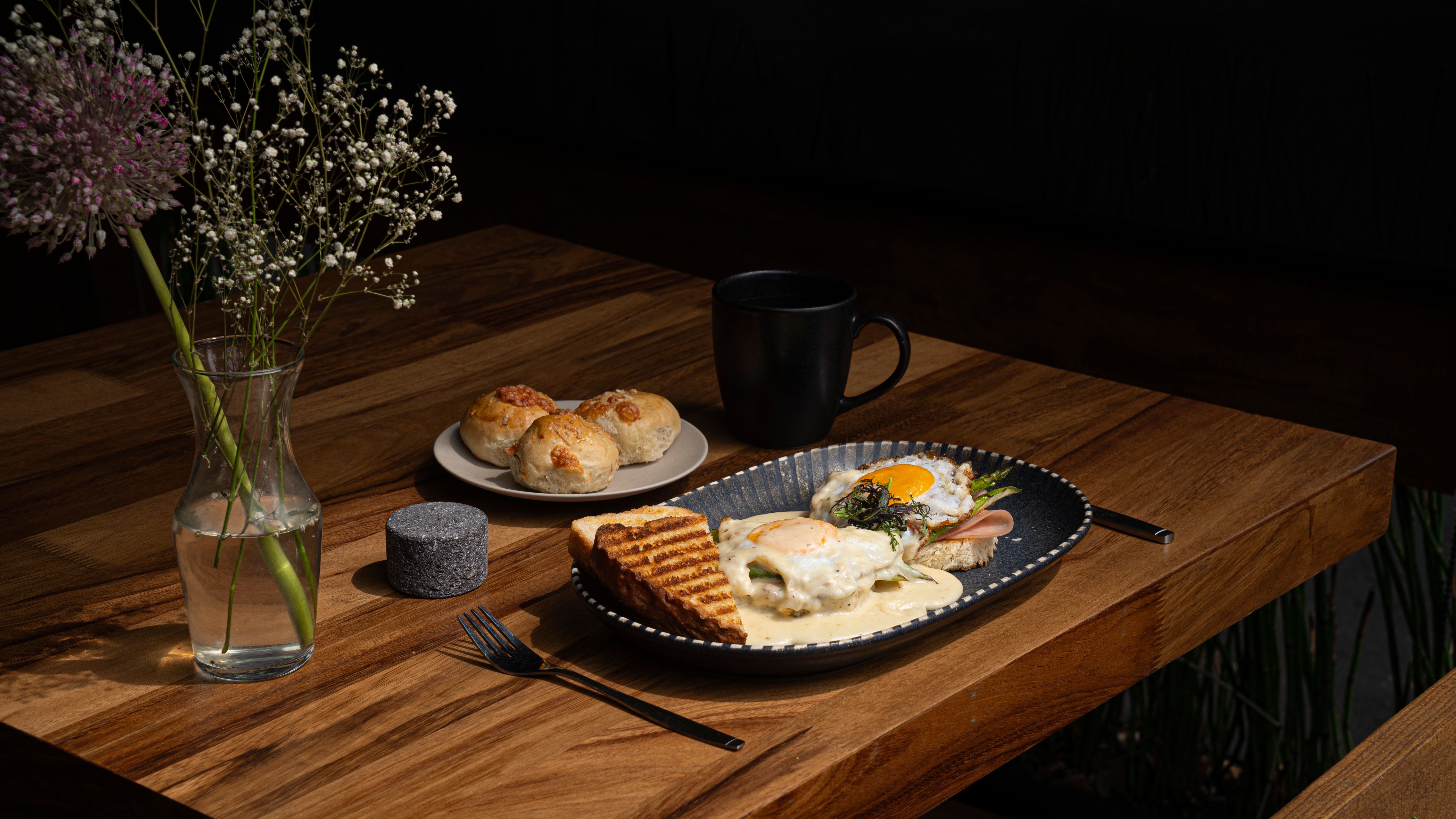 A sunlit brunch spread with eggs, greens, pastries, and coffee on a rustic table surrounded by plants.