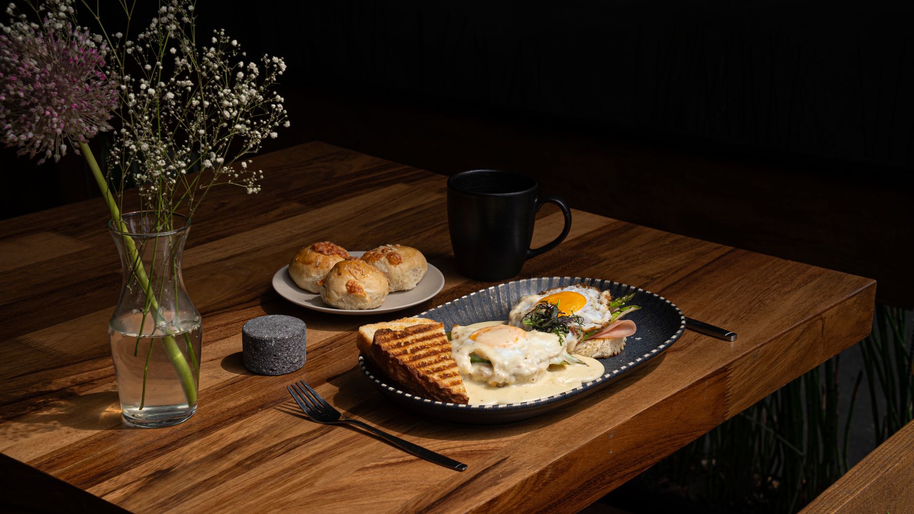 A sunlit brunch spread with eggs, greens, pastries, and coffee on a rustic table surrounded by plants.