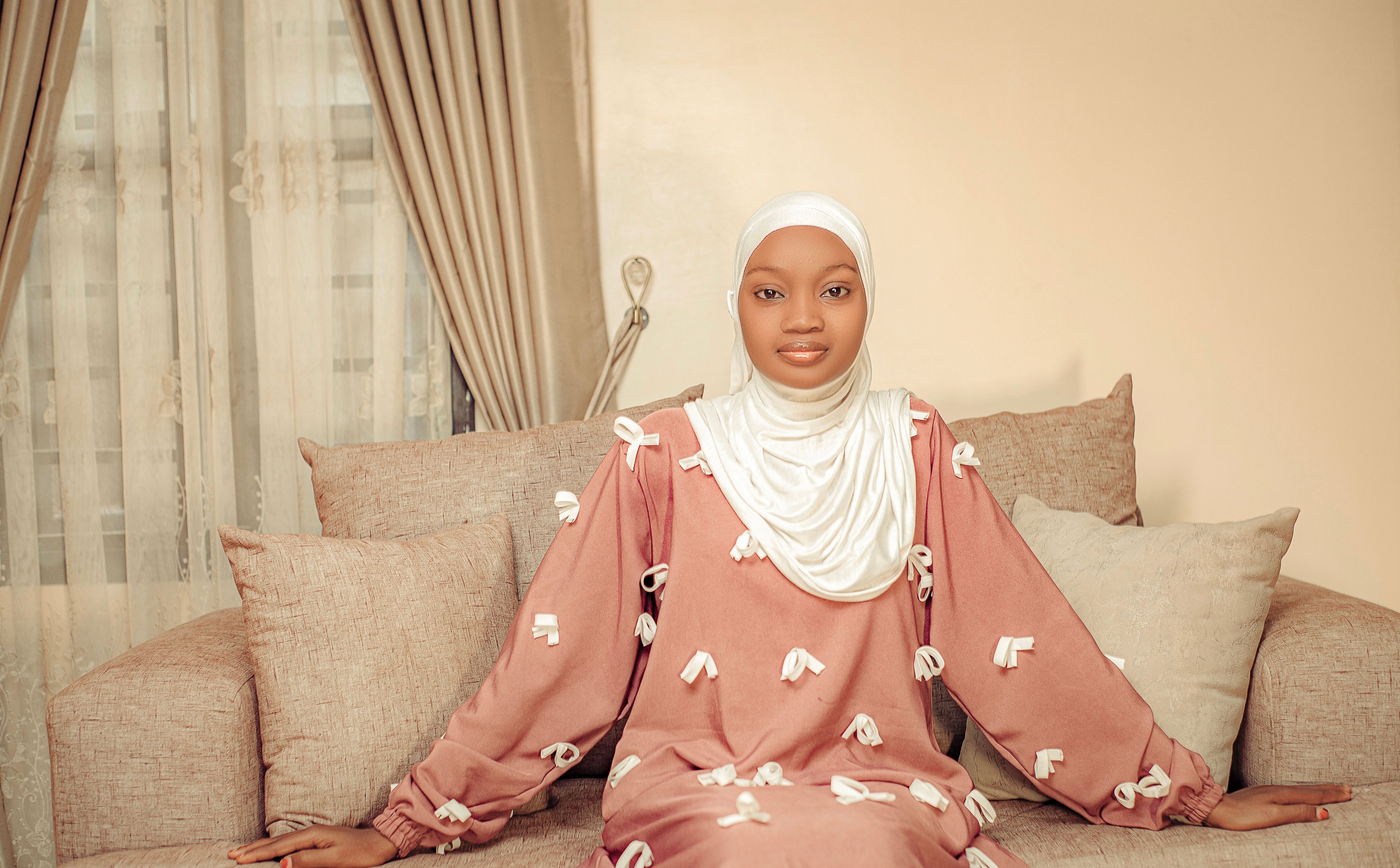 A young woman in a hijab sits elegantly on a sofa with soft lighting and neutral tones.
