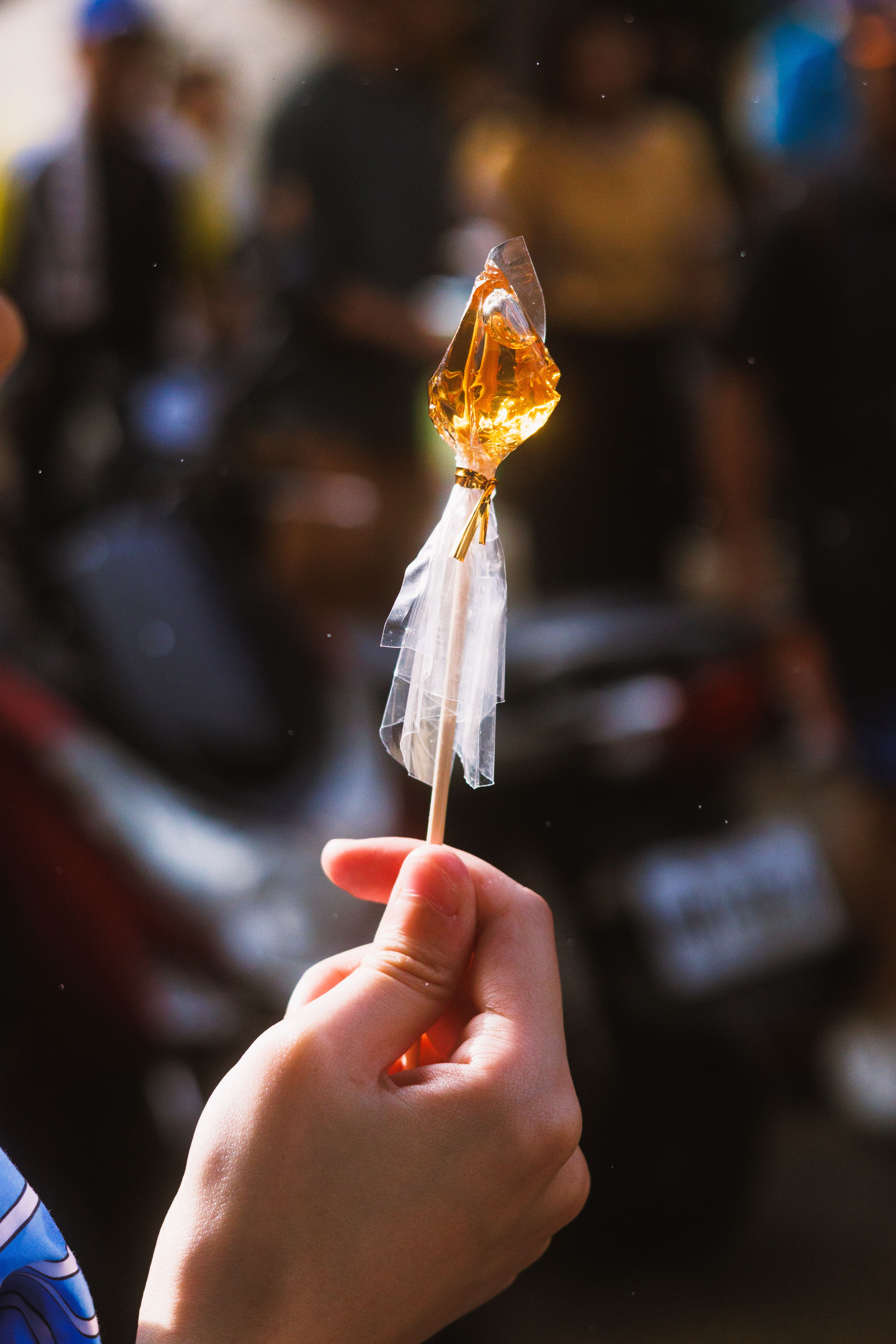A hand holds a translucent crystal sugar candy on a stick against a blurred outdoor background.