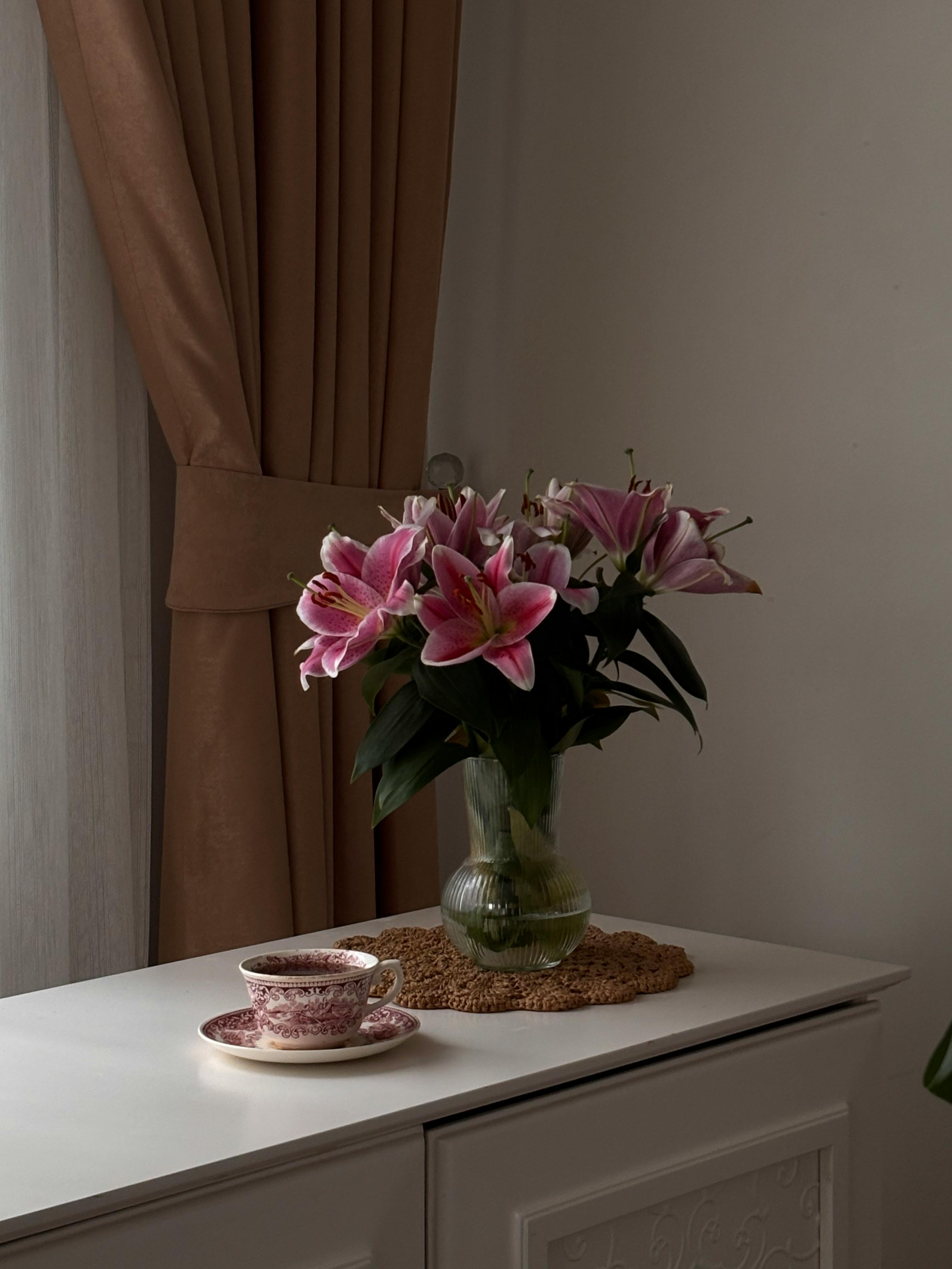 Gratuit O vază cu crini roz vibranți împodobește o masă lângă o ceașcă de ceai, într-un cadru interior senin. Fotografie de stoc