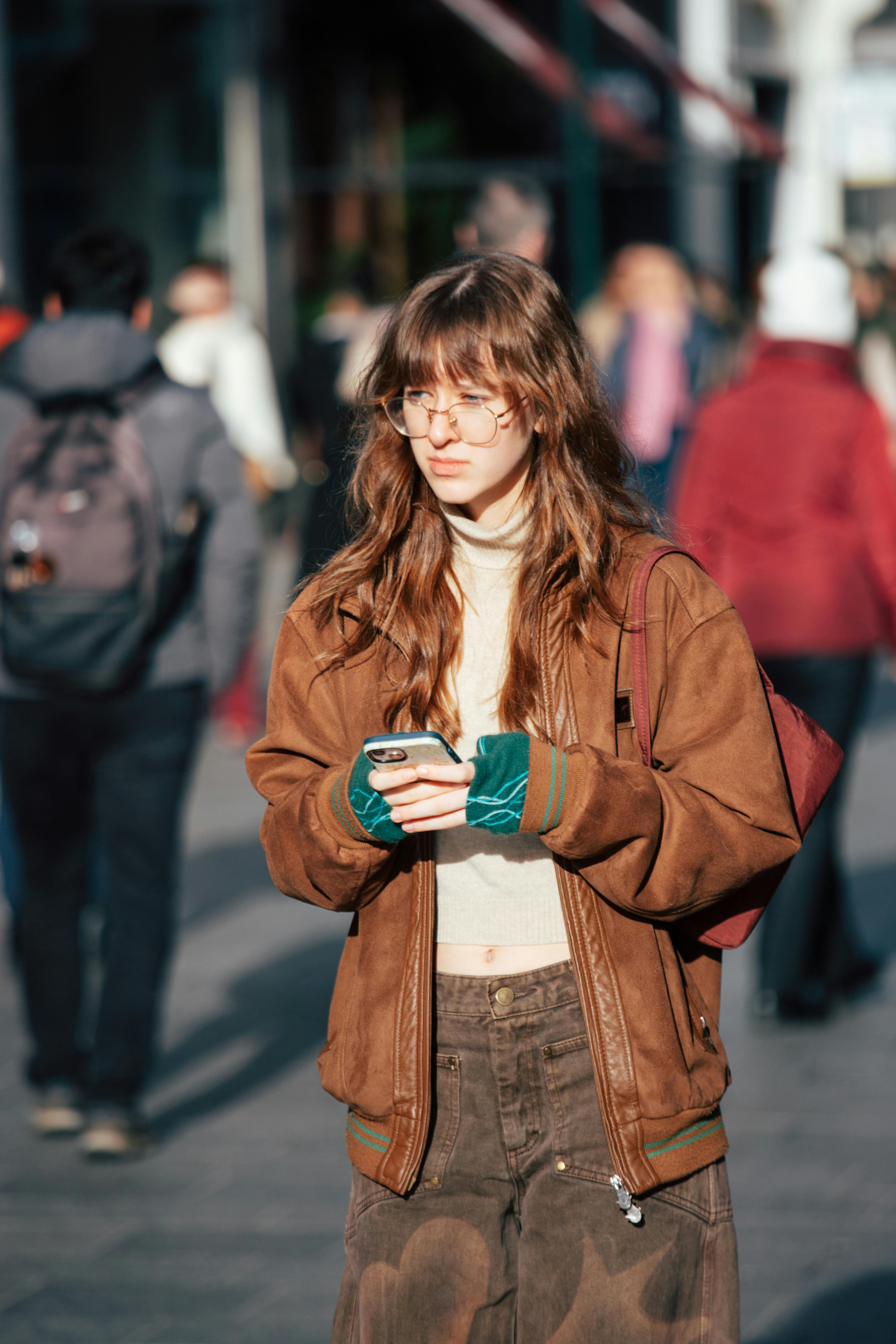 Ücretsiz Arka planda insanların olduğu bir şehir caddesinde, dışarıda akıllı telefon kullanan genç bir kadın. Stok Fotoğraflar