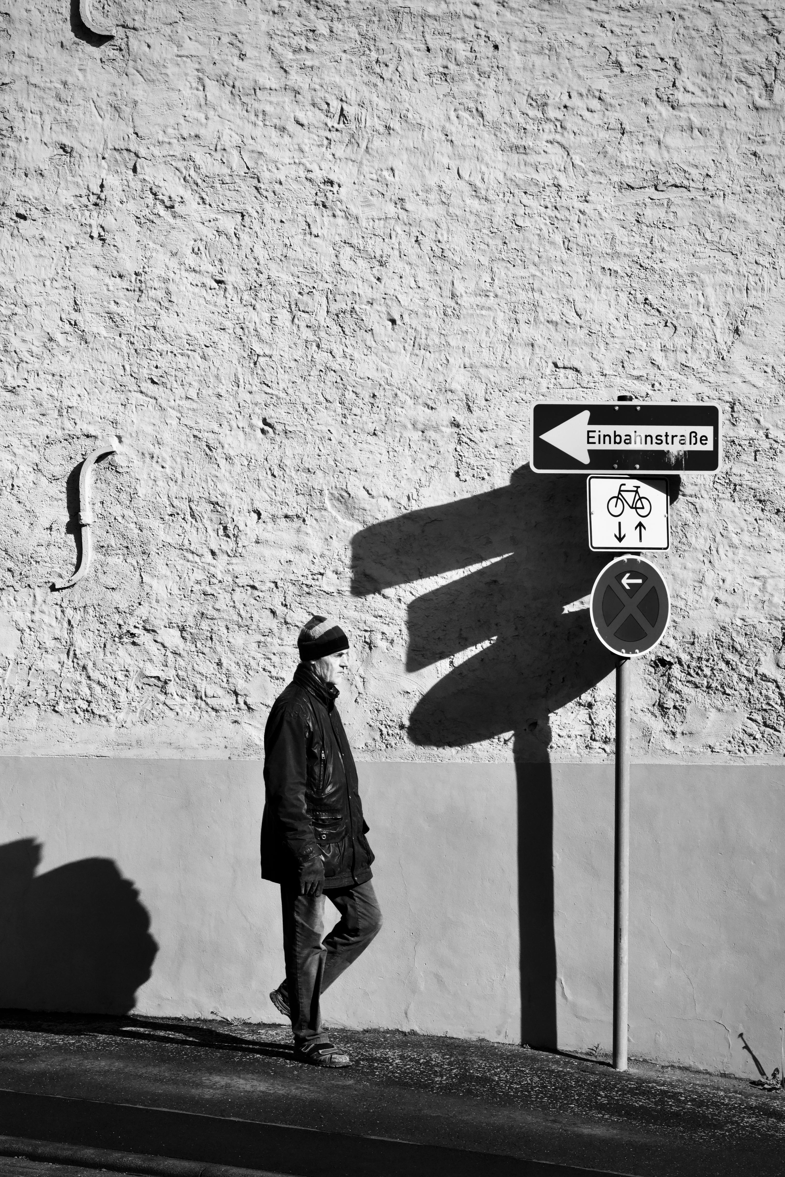 ฟรี ภาพขาวดำของคนเดินผ่านป้ายบอกทางที่ทอดเงาลงบนกำแพง คลังภาพถ่าย