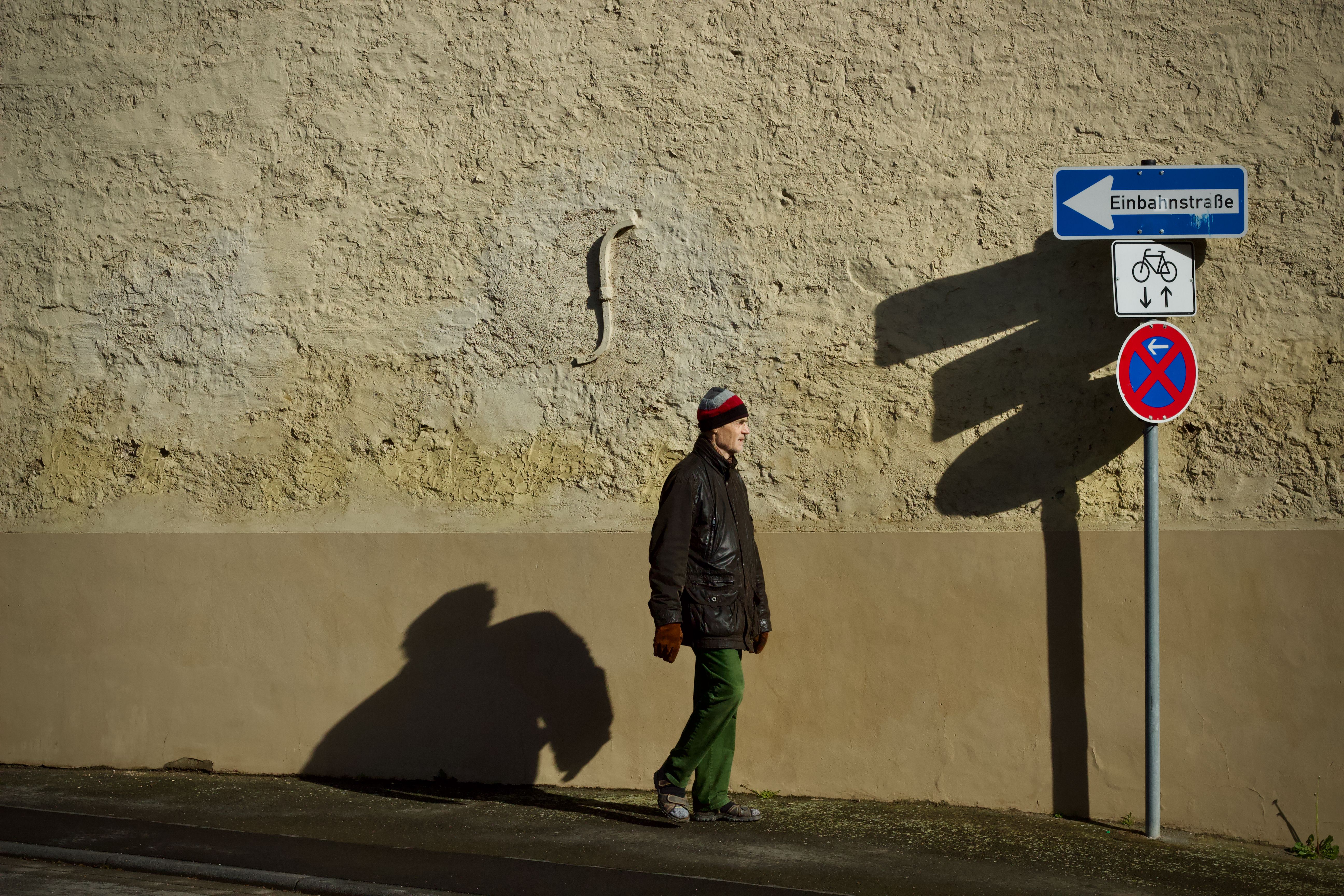Man walks by wall under street signs casting shadows in urban setting.