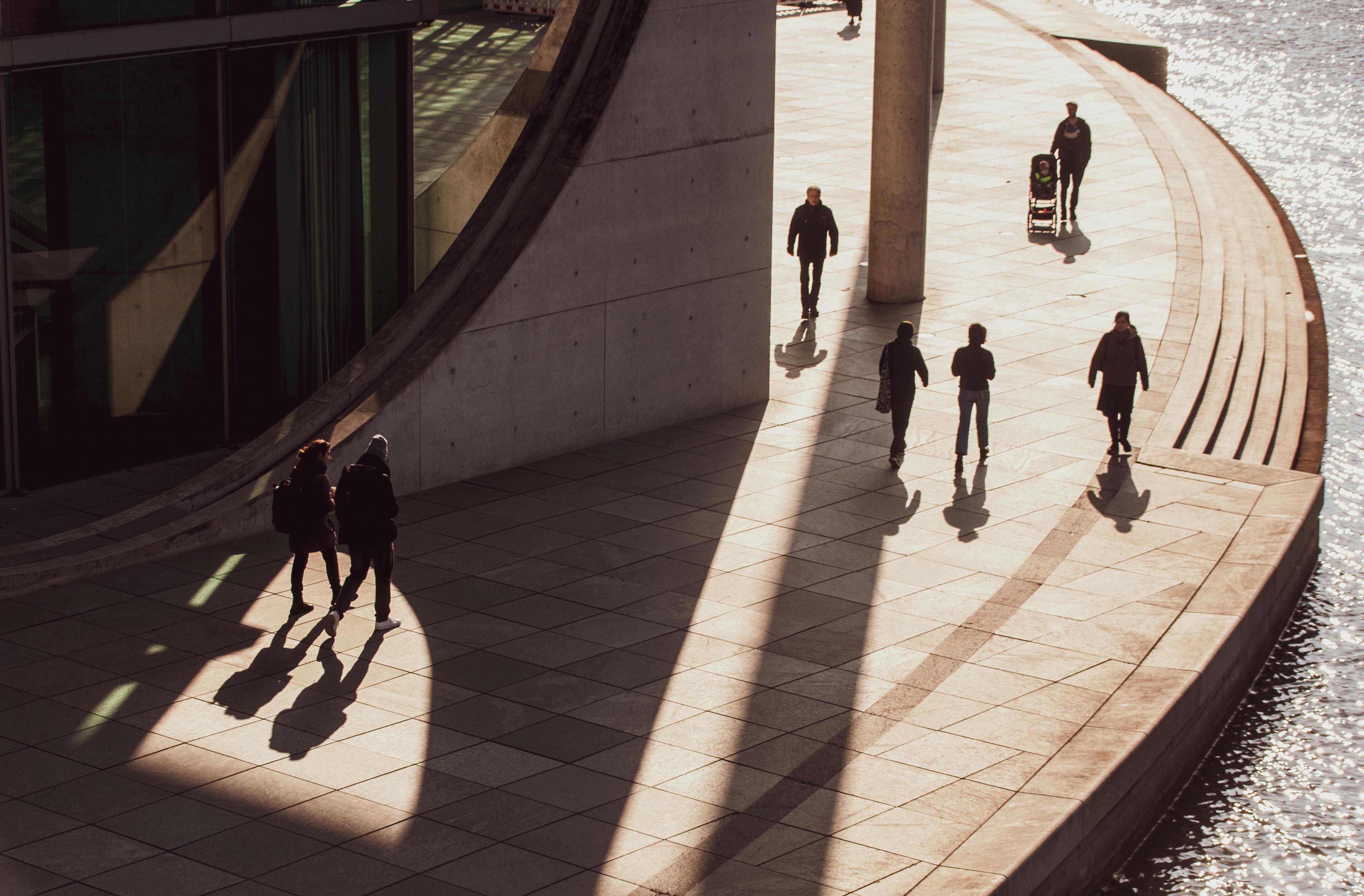 Free Silhouetted pedestrians walk along a modern riverside plaza with striking shadows. Stock Photo