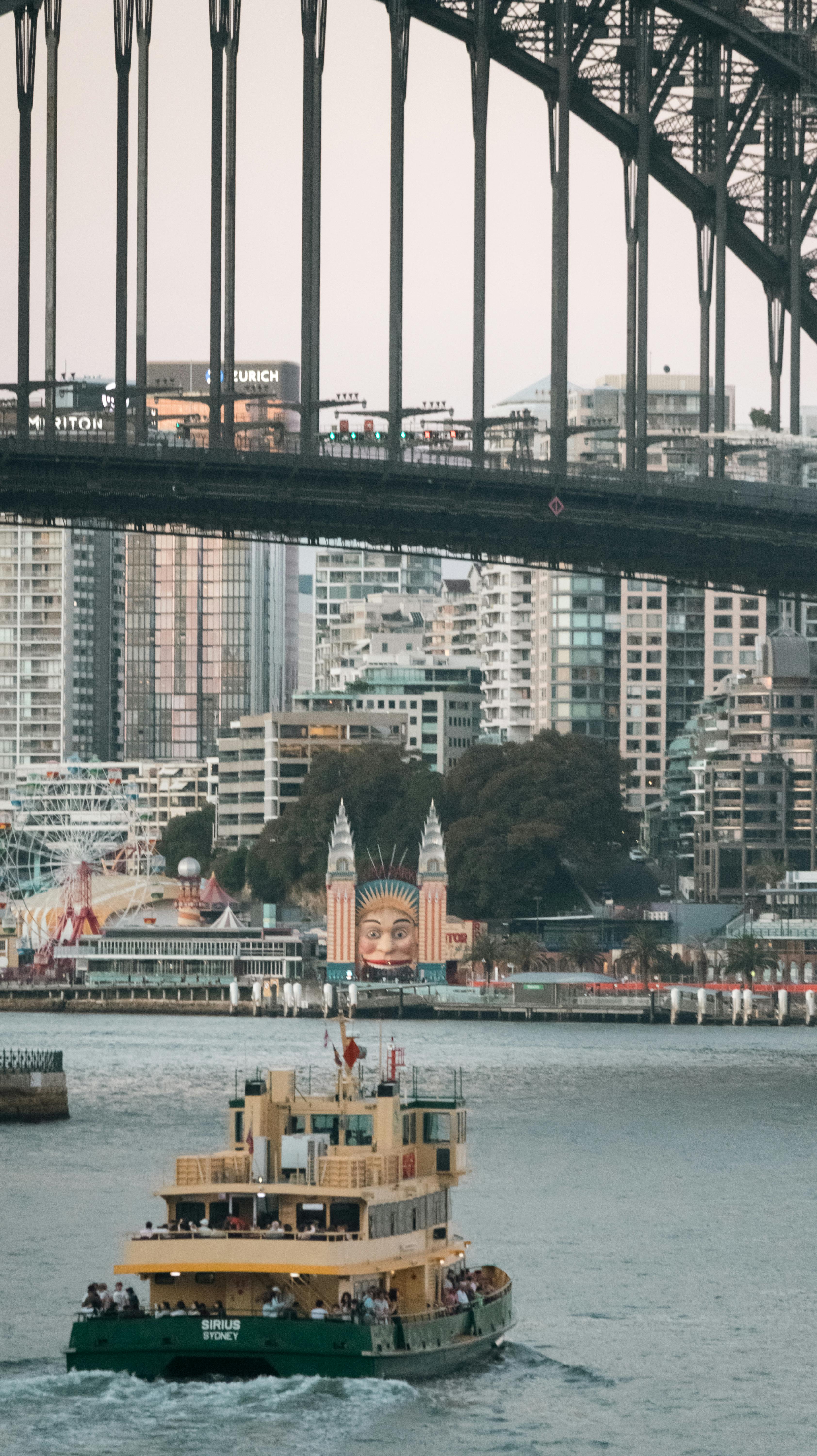 Gratuit Un ferry passe sous le pont du port de Sydney, avec Luna Park et la silhouette de la ville en arrière-plan. Photos