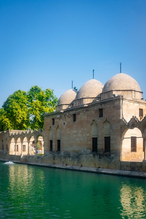 Free A beautiful historical stone building with domes by a tranquil river under a clear blue sky. Stock Photo