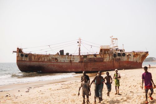 Free Rusty shipwreck on a sunny beach in Freetown with children playing nearby. Stock Photo