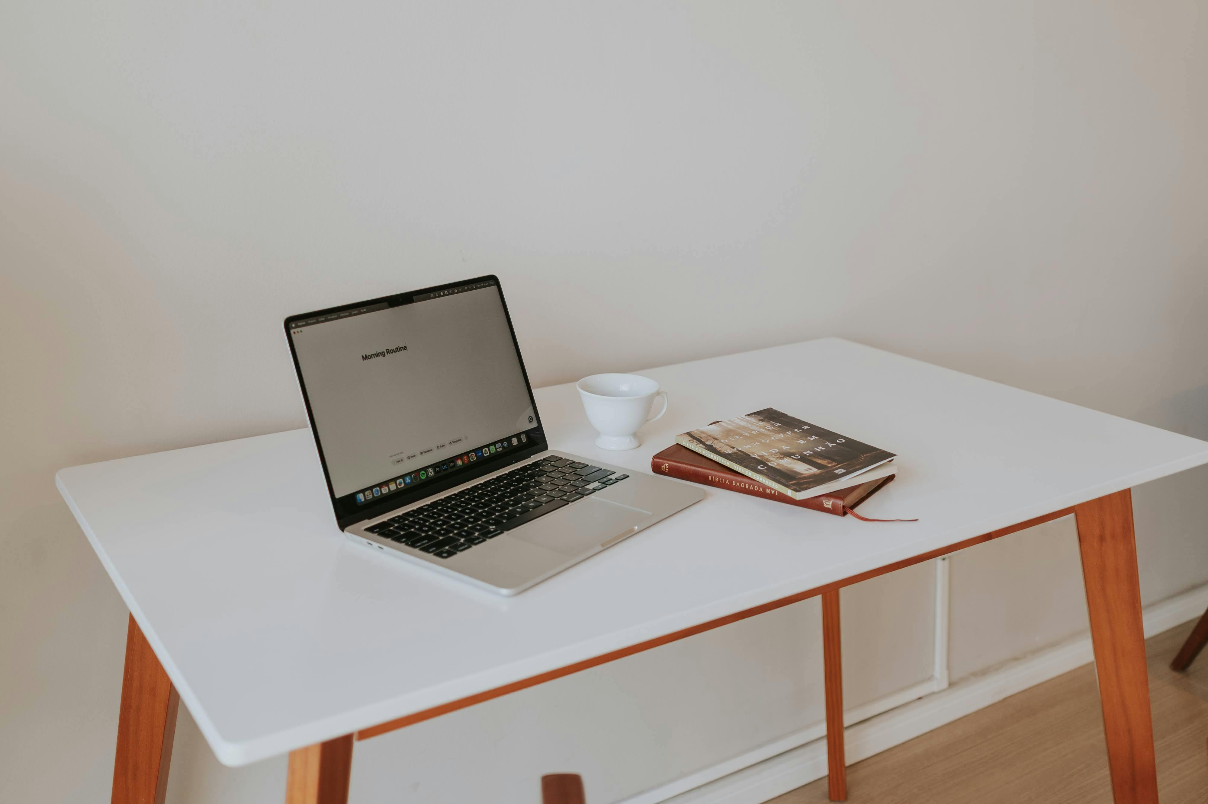 A minimalistic workspace featuring a laptop, coffee cup, and books on a white desk with wooden legs.