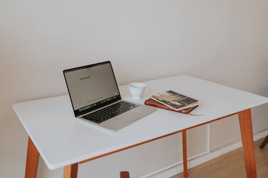 A minimalistic workspace featuring a laptop, coffee cup, and books on a white desk with wooden legs.