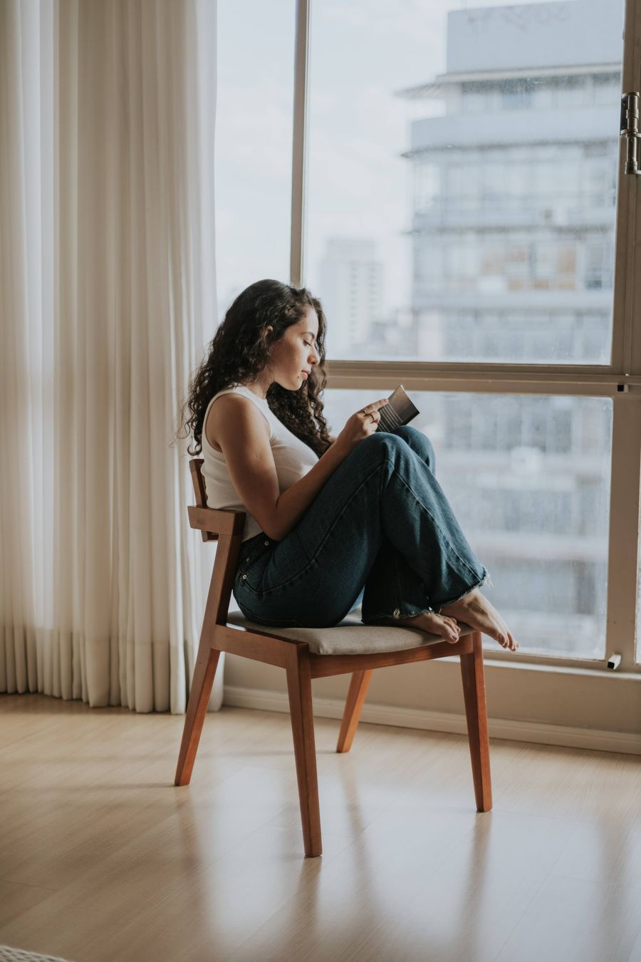 A woman reading on a chair by a tall apartment window, feet tucked up, on a quiet afternoon