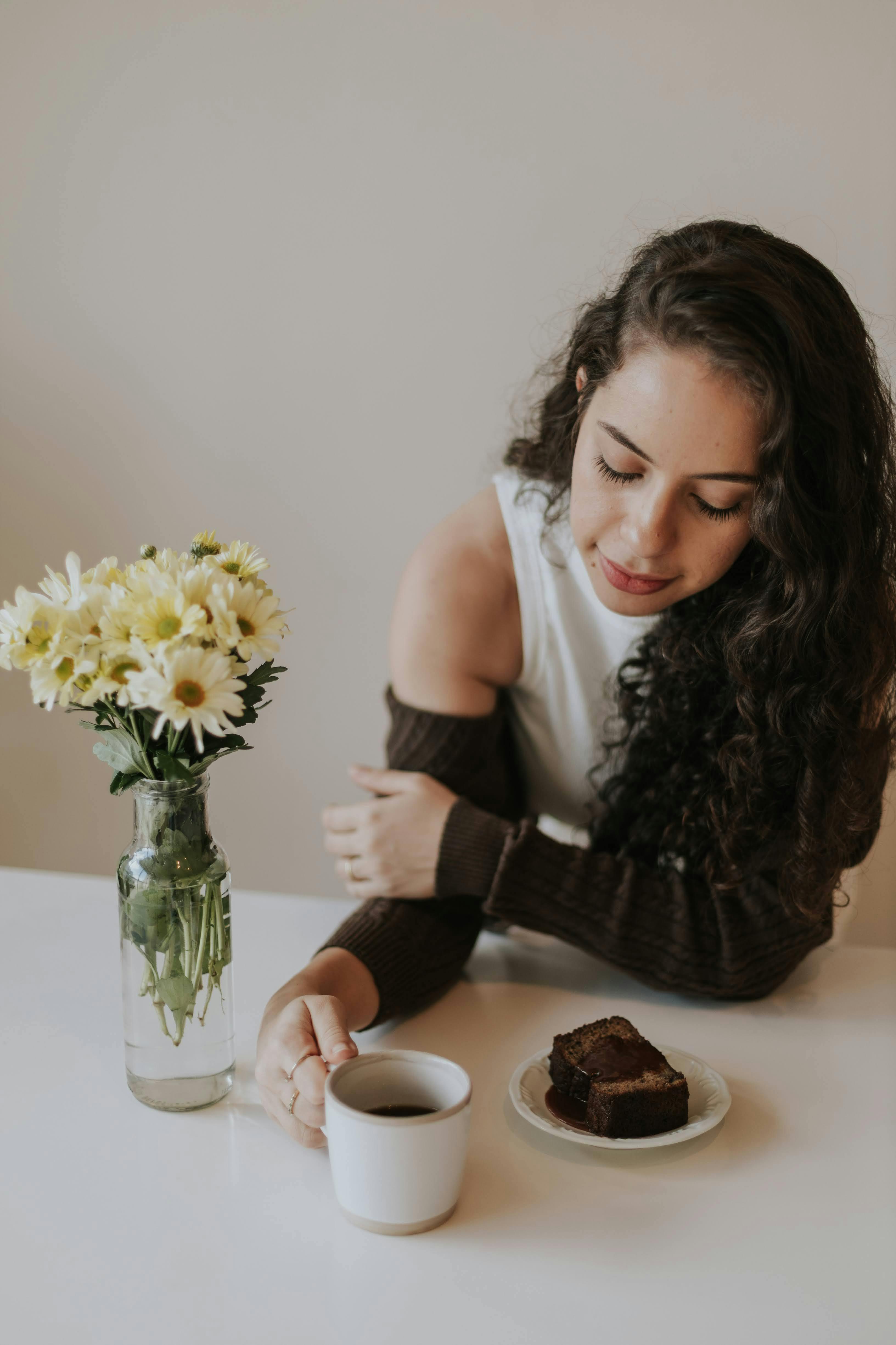 Gratis Una giovane donna con i capelli ricci si gode un caffè e una fetta di torta seduta a un tavolo decorato con fiori, creando un'atmosfera accogliente. Foto a disposizione