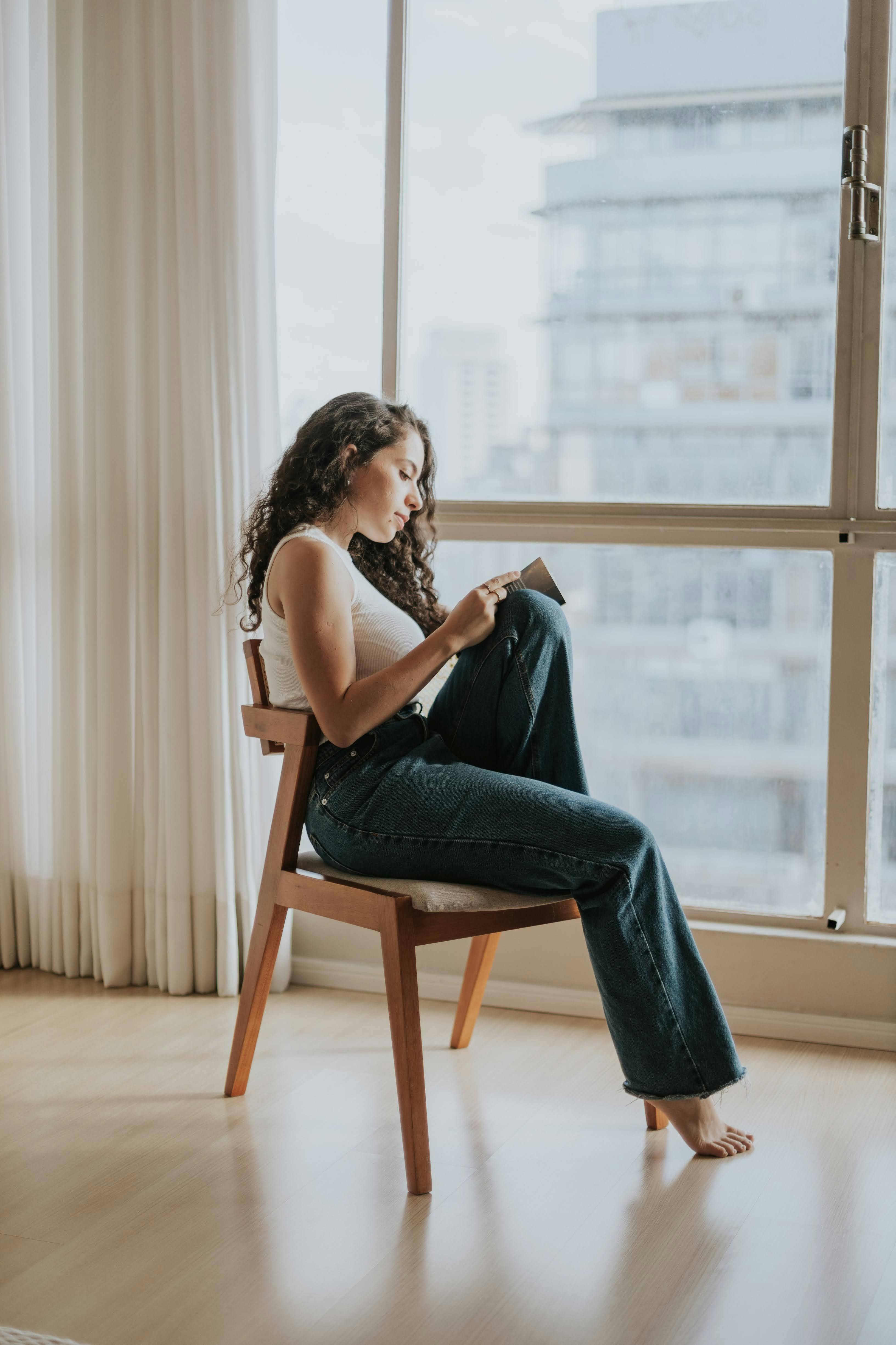 ฟรี หญิงสาวนั่งพักผ่อนอ่านหนังสืออยู่ริมหน้าต่างบานใหญ่ สร้างบรรยากาศที่เงียบสงบภายในบ้าน คลังภาพถ่าย