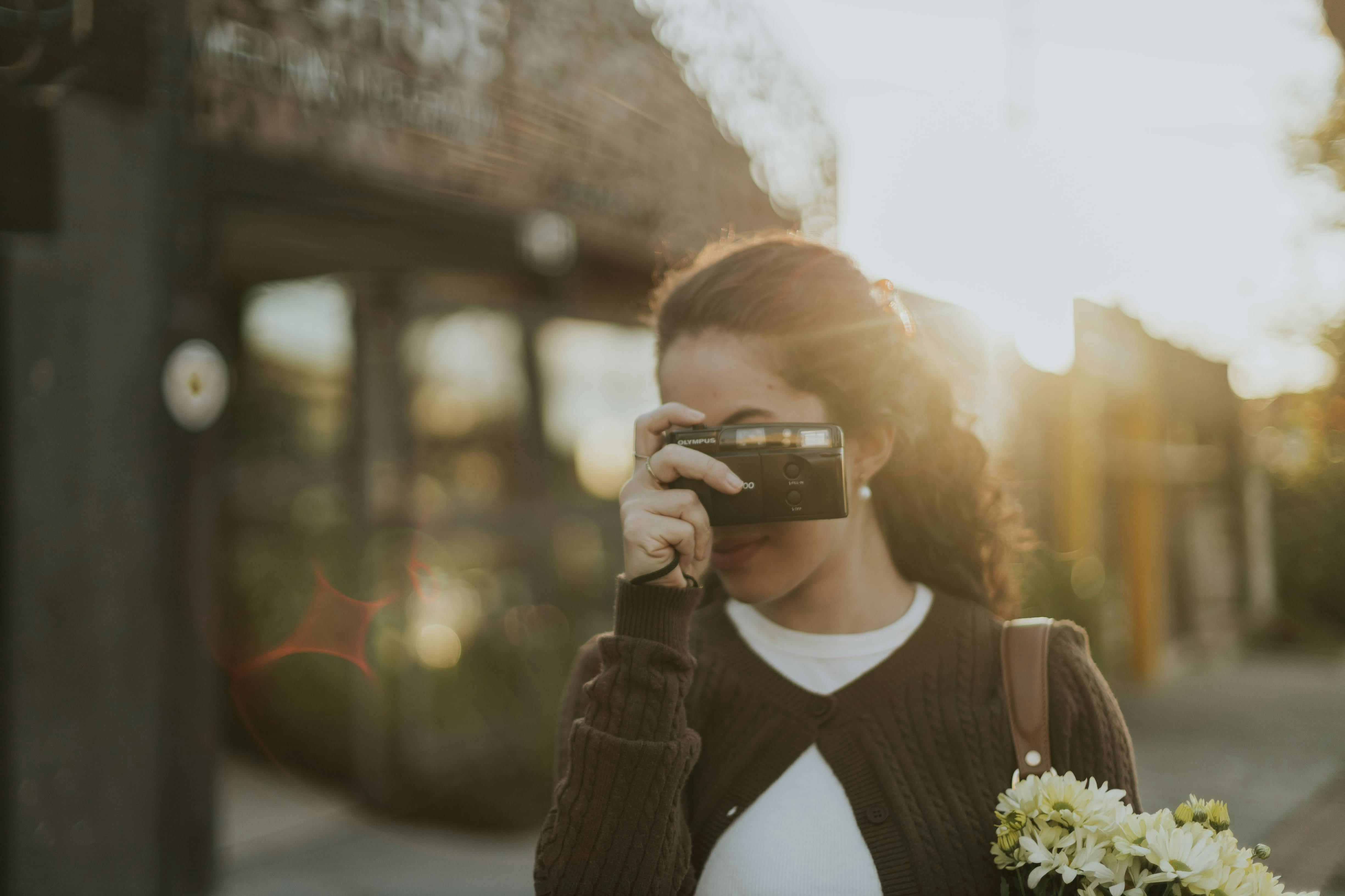 bezplatná Mladá žena fotí venku historickým fotoaparátem při západu slunce a drží květiny. Základová fotografie