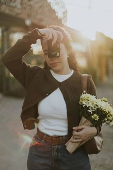 Young woman capturing a moment with a vintage camera, holding flowers, in warm sunlight.