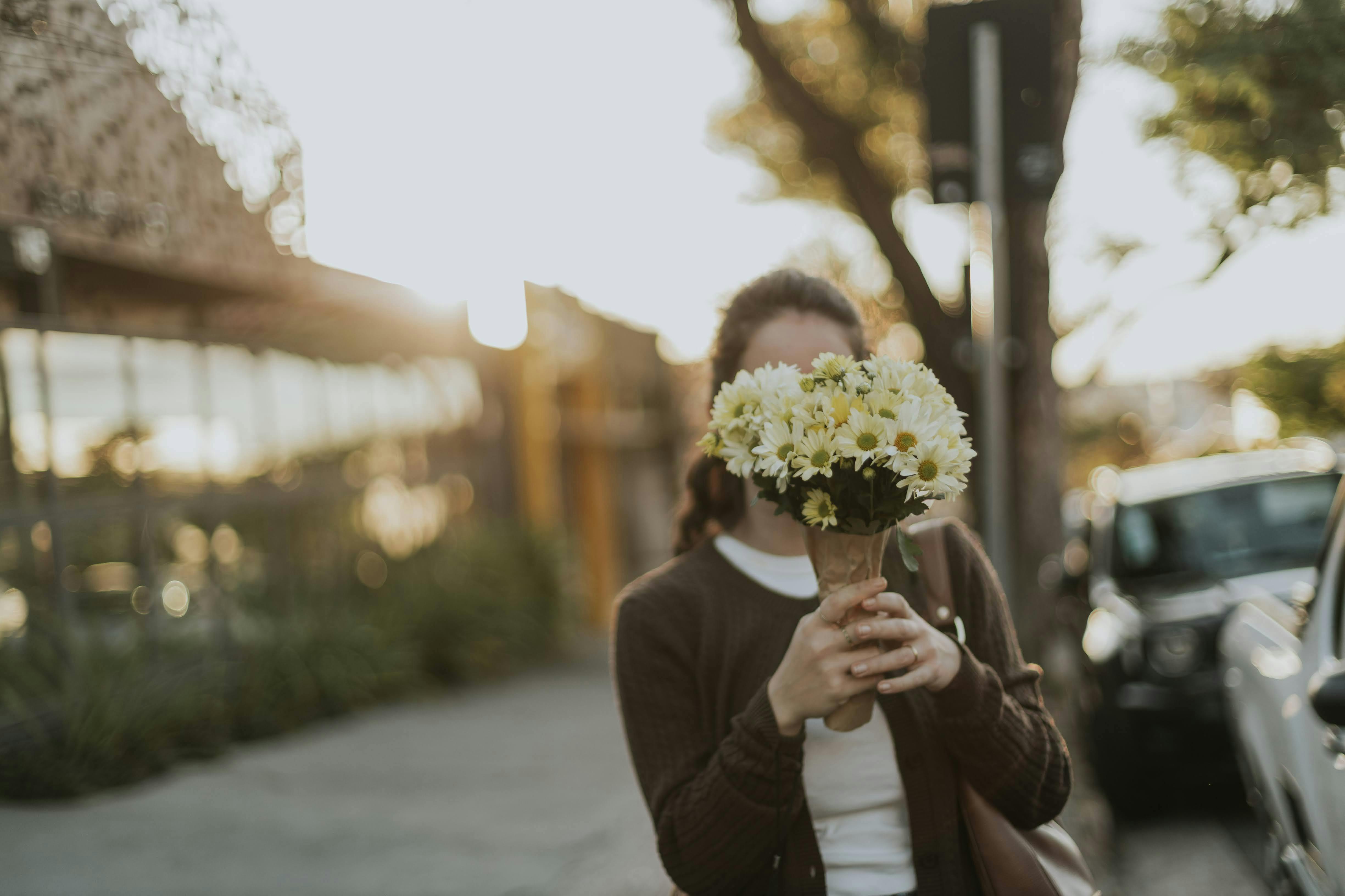 Bezpłatne Kobieta zakrywająca twarz bukietem żółtych i białych kwiatów na oświetlonej słońcem ulicy. Zdjęcie z galerii