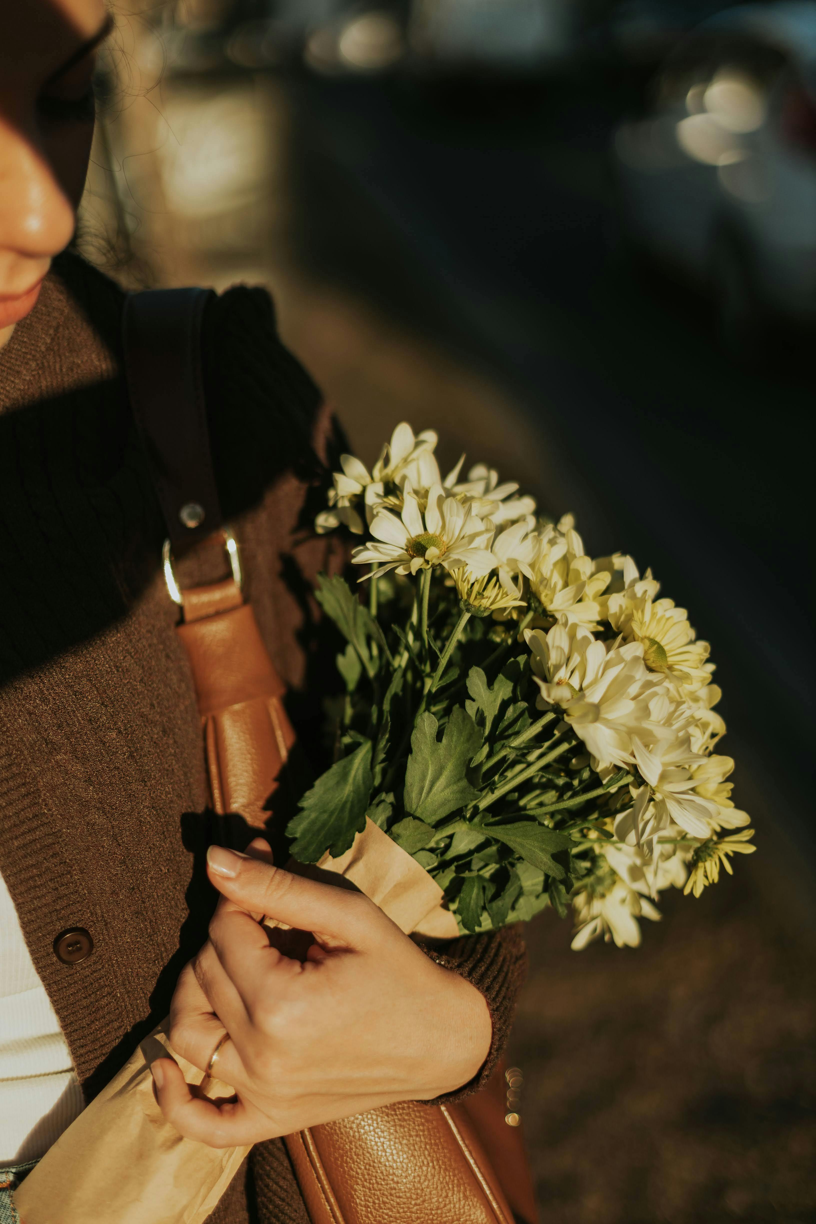 Gratis Una donna adulta tiene in mano un mazzo di margherite bianche all'aperto, nella calda luce del tardo pomeriggio, creando un'atmosfera serena e riflessiva. Foto a disposizione