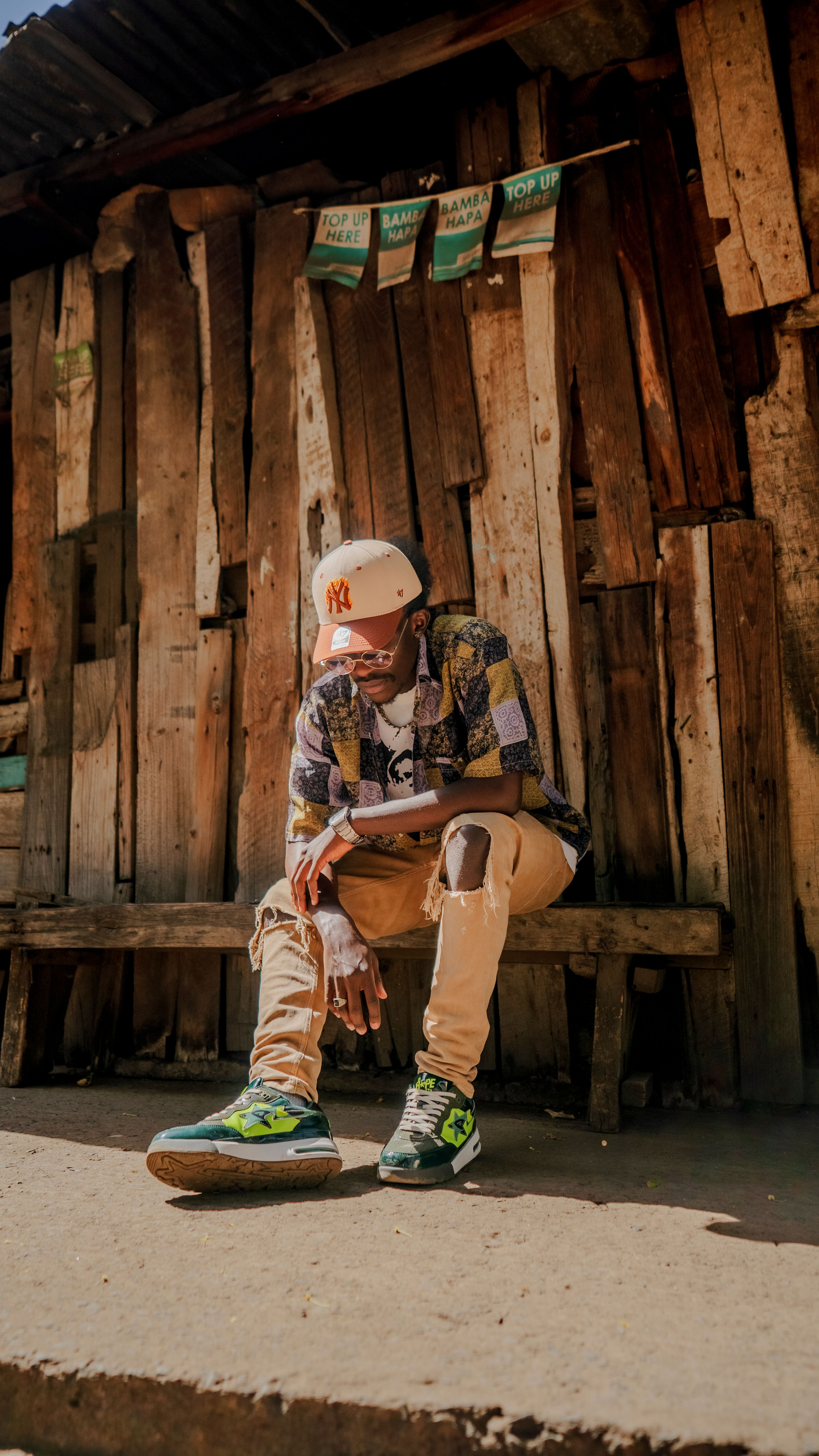 A young man in vibrant attire sits outside a rustic wooden structure in Nairobi, Kenya, exuding cool confidence.