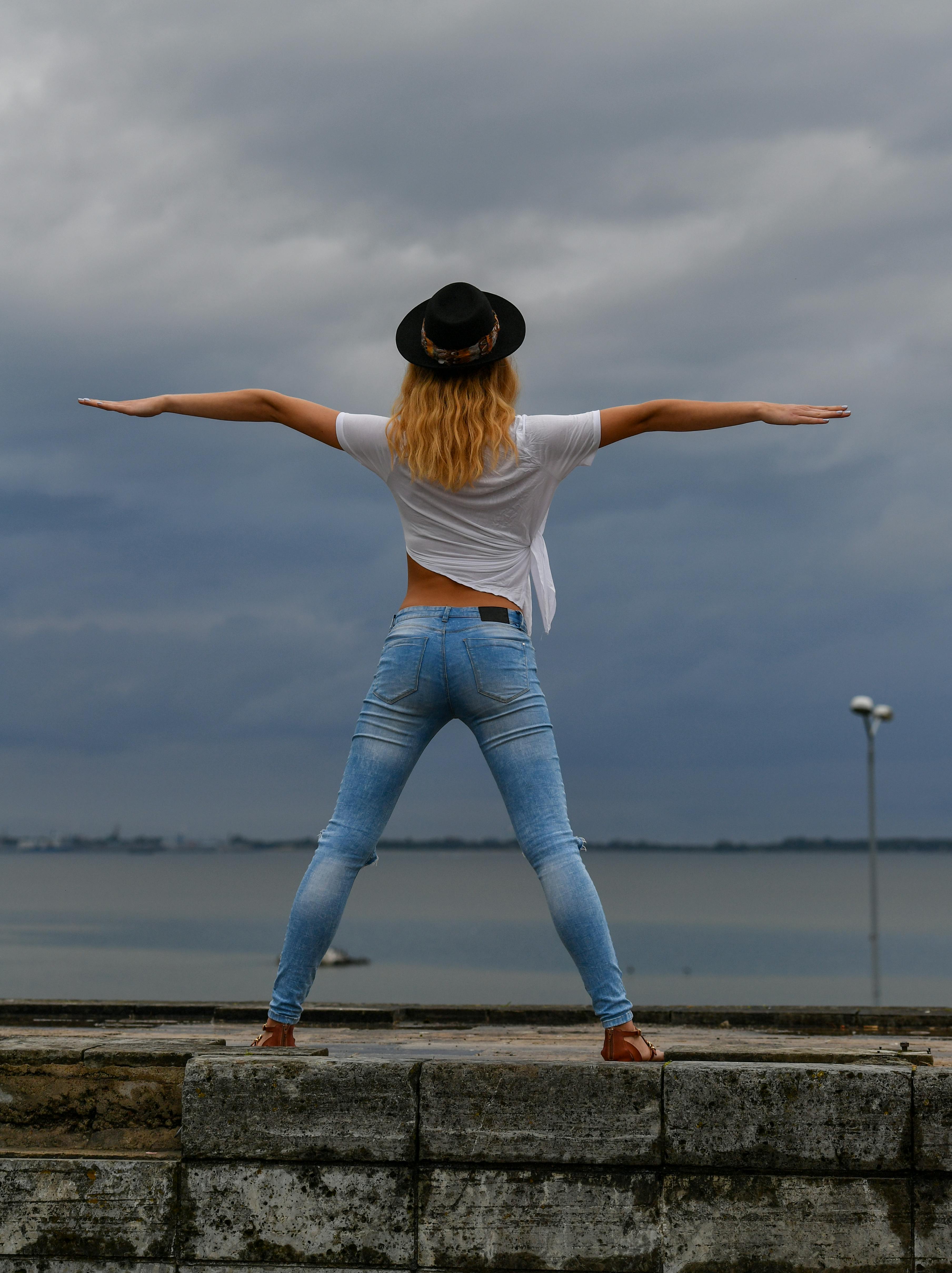 ฟรี หญิงสาวผมบลอนด์สวมกางเกงยีนส์และหมวก โพสท่าริมทะเลโดยมีฉากหลังเป็นท้องฟ้าที่สวยงาม แสดงให้เห็นถึงวิถีชีวิตและแฟชั่นสมัยใหม่ คลังภาพถ่าย