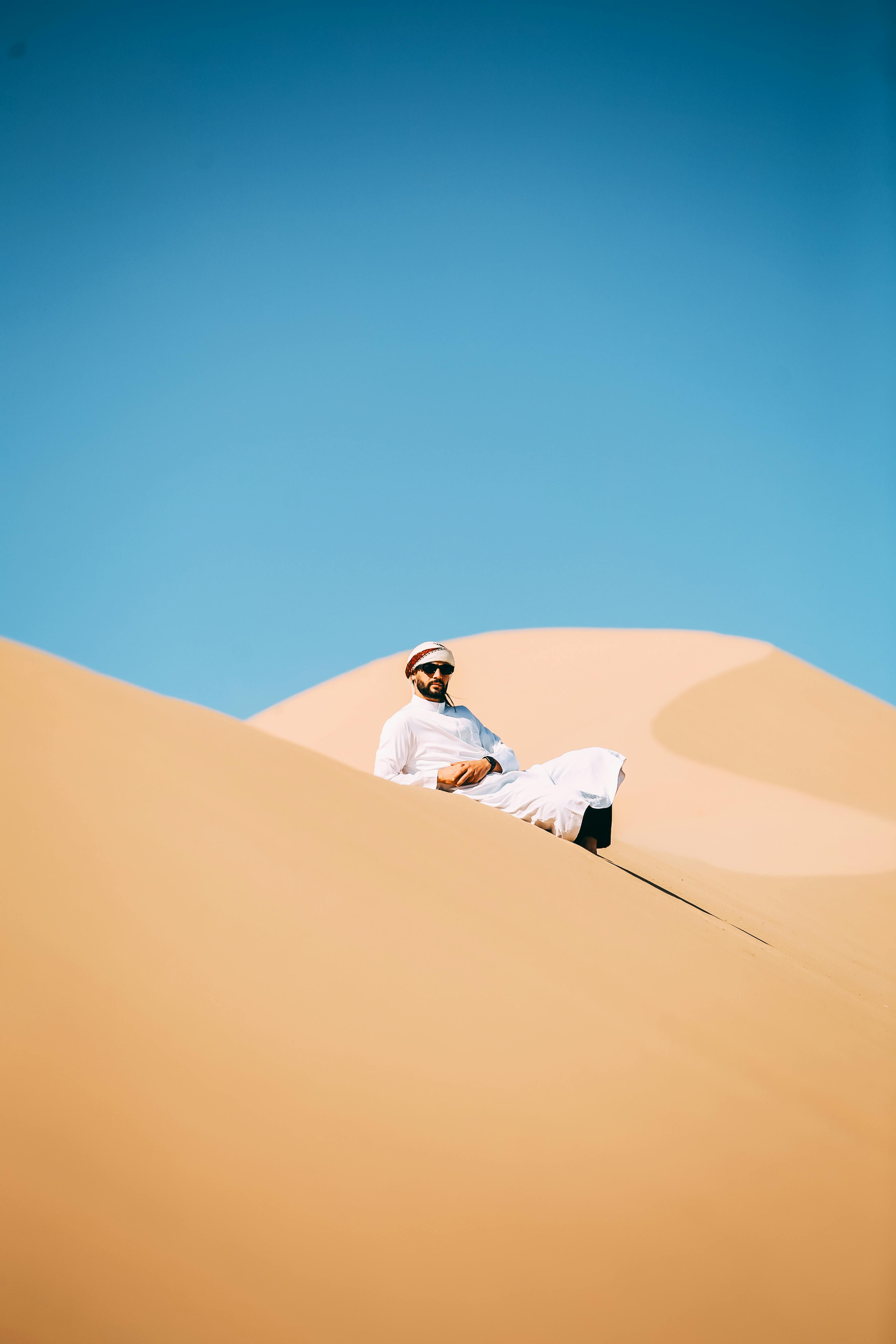 Gratis Un uomo in abiti tradizionali si rilassa sulle dune di sabbia illuminate dal sole, contro un cielo azzurro e terso. Foto a disposizione