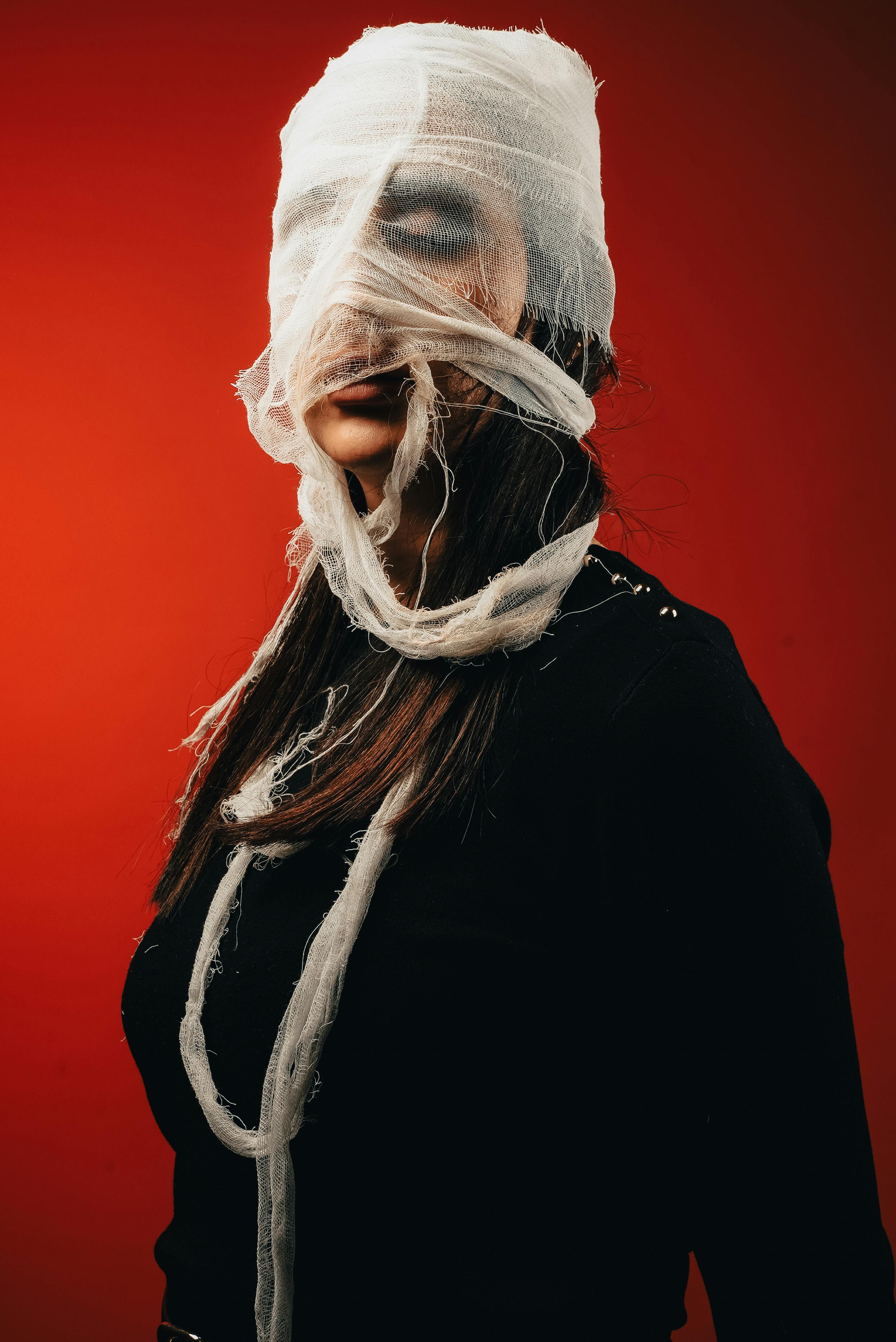 Free Dramatic portrait of a woman with face obscured by bandages against a bold red background. Stock Photo