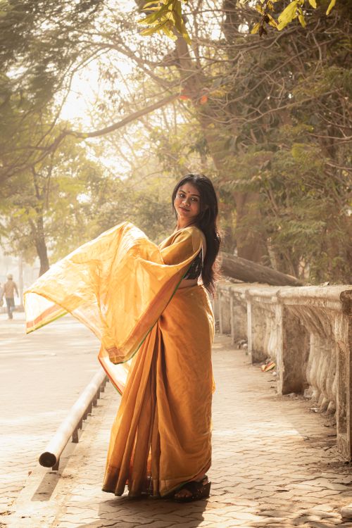Free A woman in a traditional saree enjoying a sunny day outdoors amidst trees and urban scenery. Stock Photo