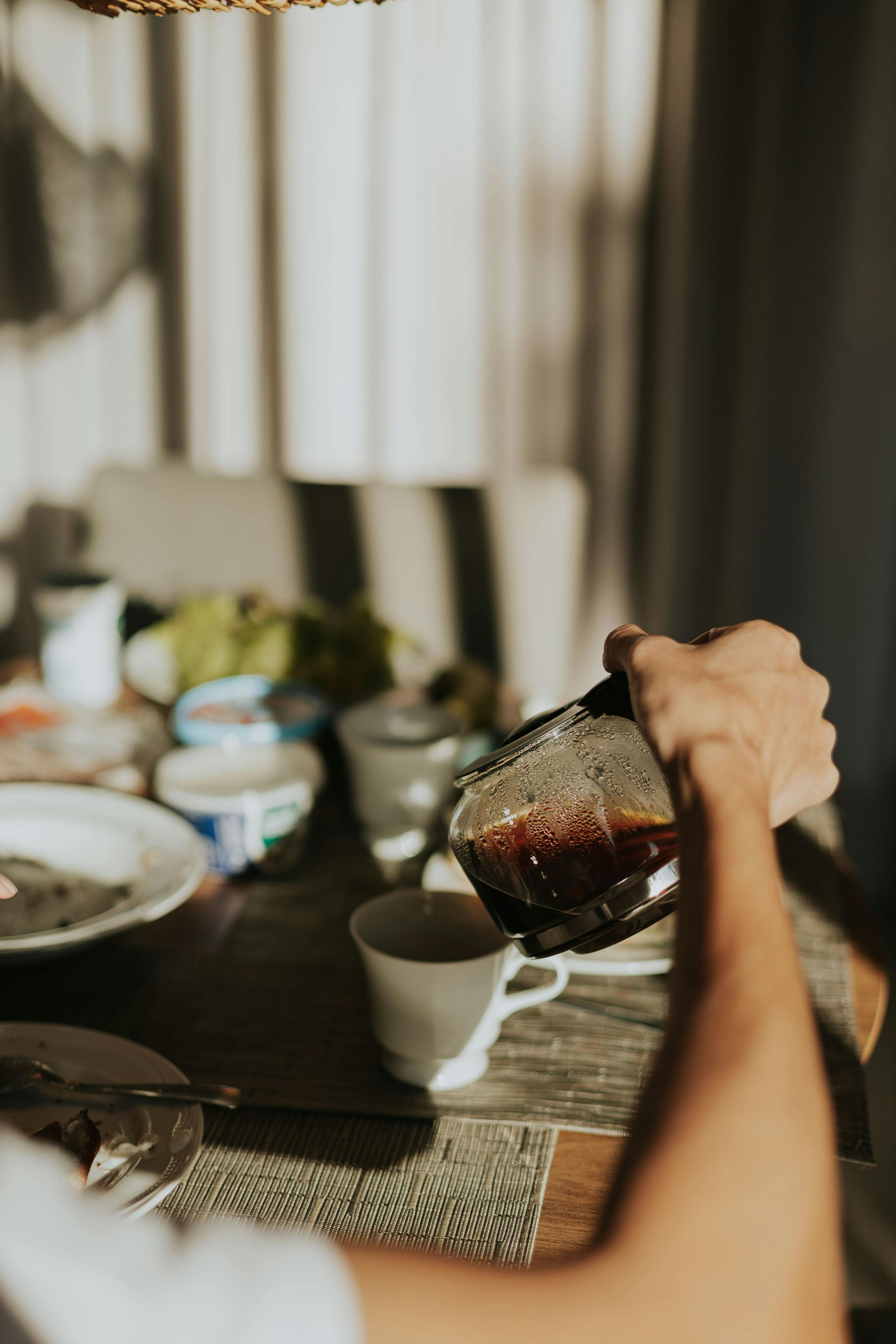 Gratis Una persona versa il caffè da una brocca di vetro in una tazza bianca in una cucina illuminata soffusamente. Foto a disposizione