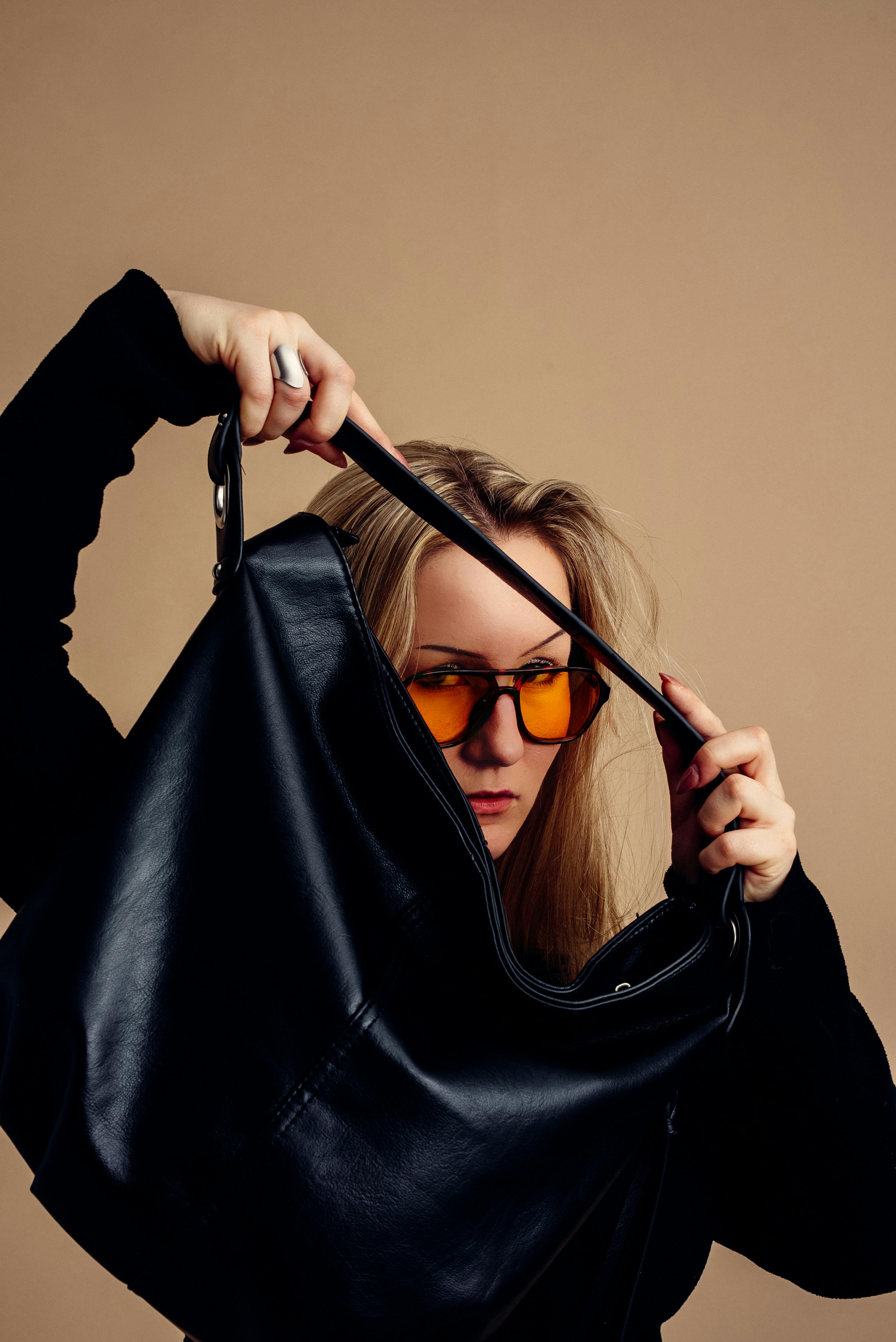 bezplatná Stylová žena se slunečními brýlemi si prohlíží černou koženou kabelku na neutrálním pozadí. Základová fotografie