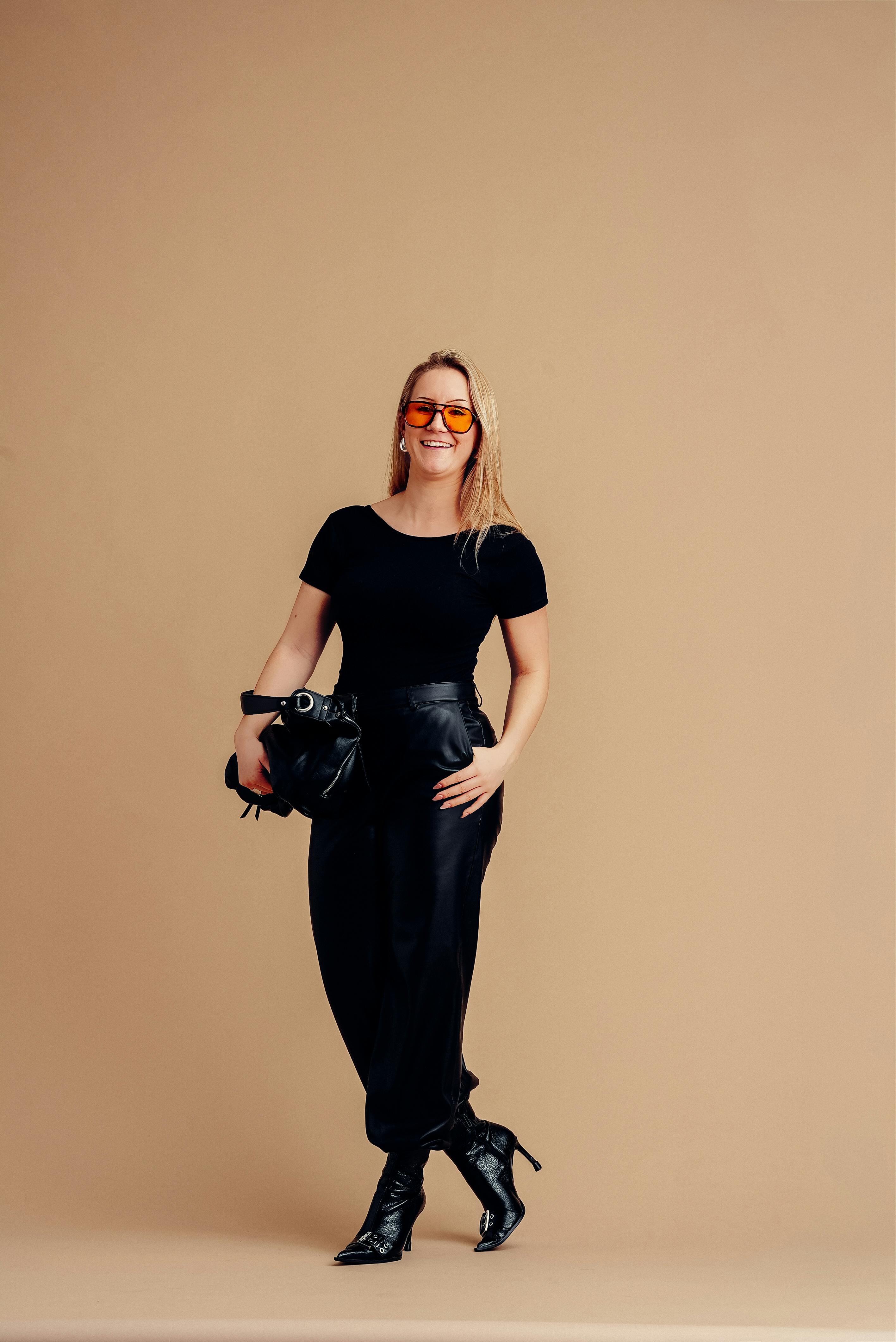 Gratis Donna alla moda in abito nero con occhiali da sole alla moda, posa con sicurezza su uno sfondo beige caldo. Foto a disposizione