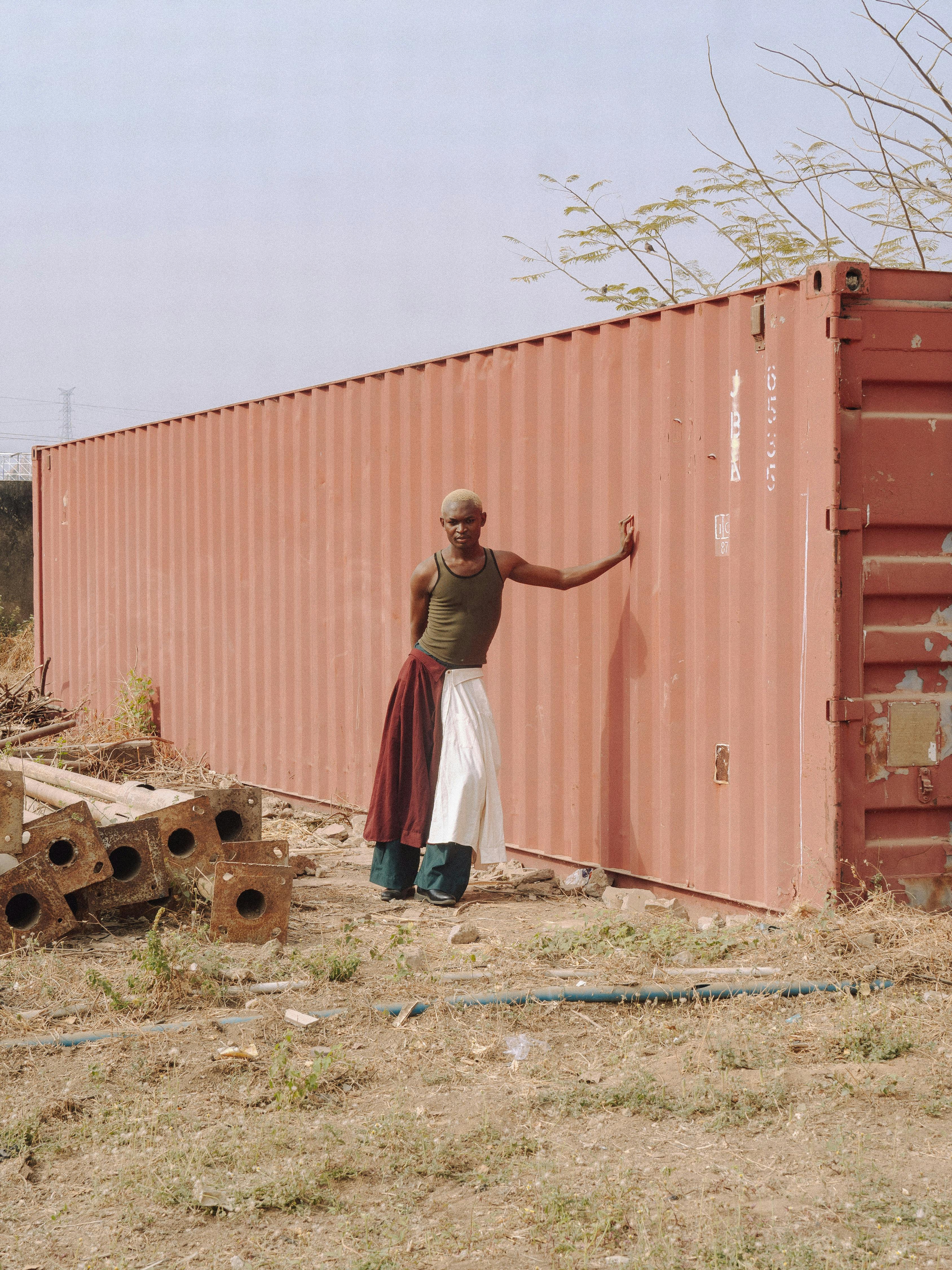 Adult man posing beside a red shipping container outdoors in urban Abuja.
