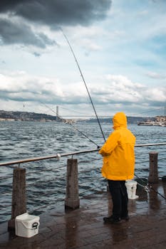 Fisherman in yellow raincoat at Bosphorus shore with cloudy sky.