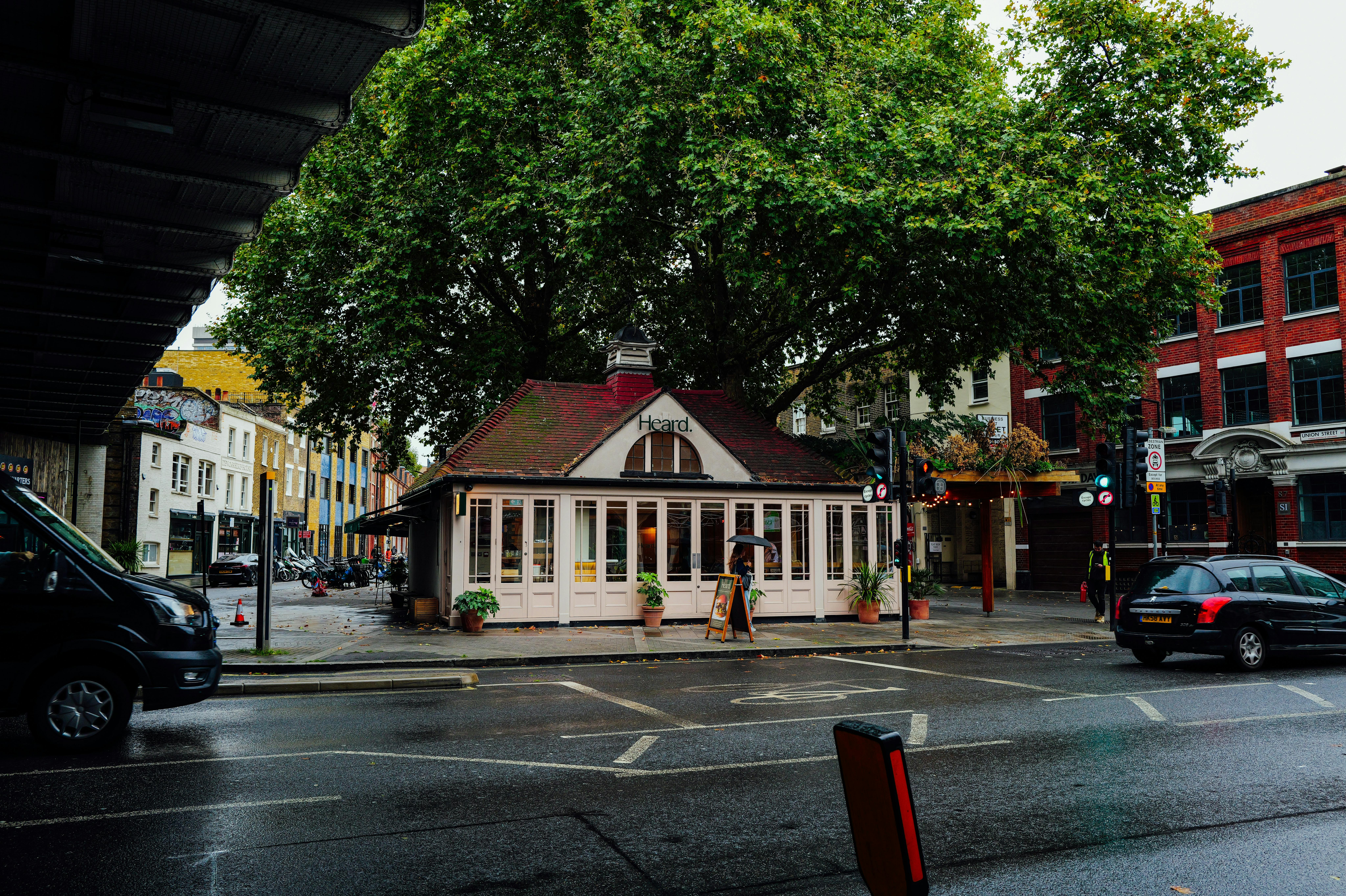 Kostenlos Ein malerisches Café schmiegt sich unter einen großen Baum an einer regnerischen Straßenecke und bietet einen gemütlichen Rückzugsort in der Stadt. Stock-Foto