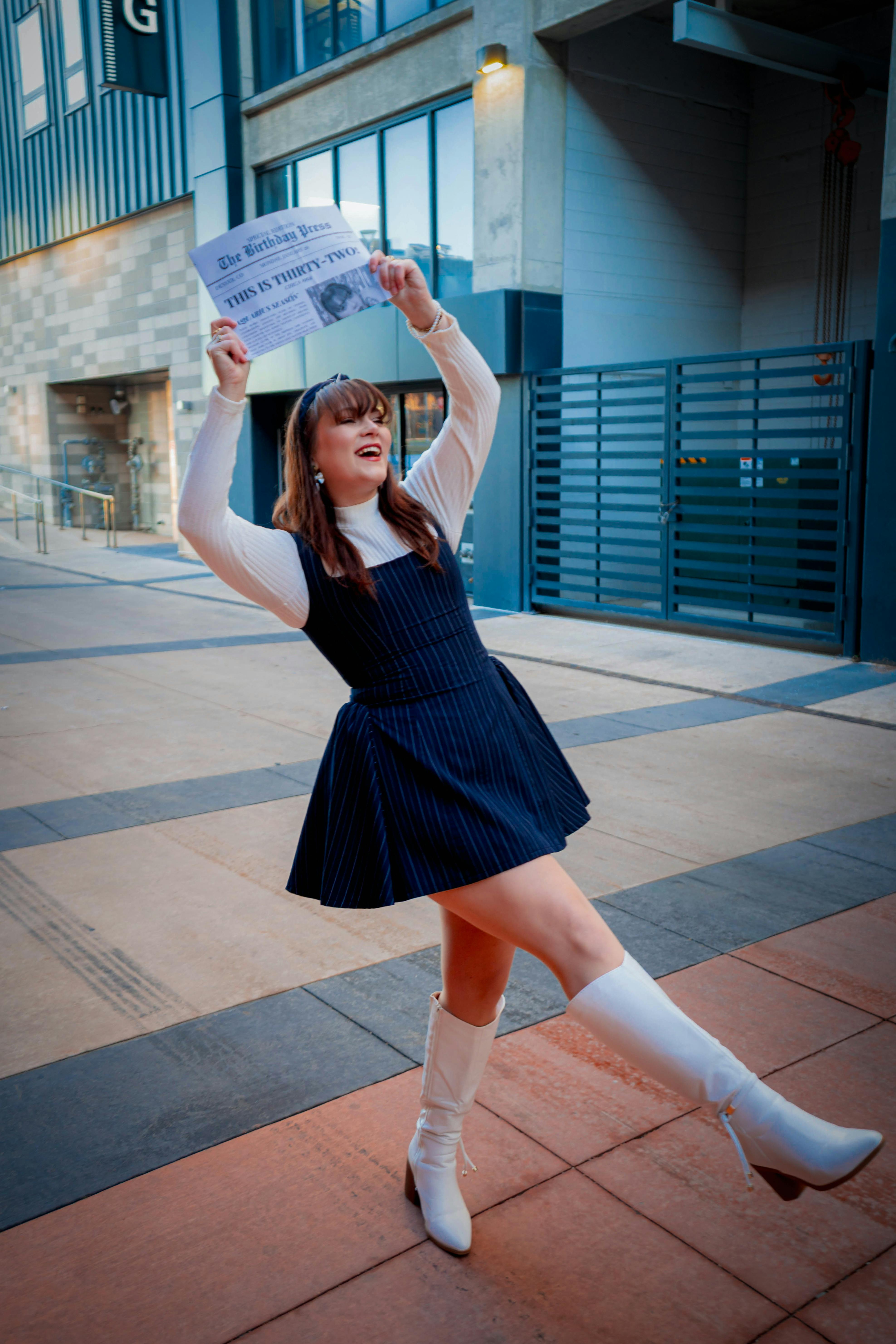 Gratis Una giovane donna elegante festeggia, tenendo in mano un giornale, in una vivace scena di strada cittadina. Foto a disposizione