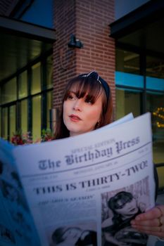 A stylish woman reads a birthday newspaper outdoors, showcasing winter fashion in an urban setting.