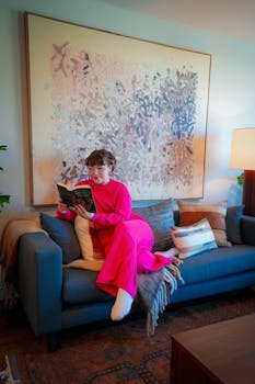 Woman in bright pink outfit reading a book on a comfortable sofa in a modern, well-decorated living room.
