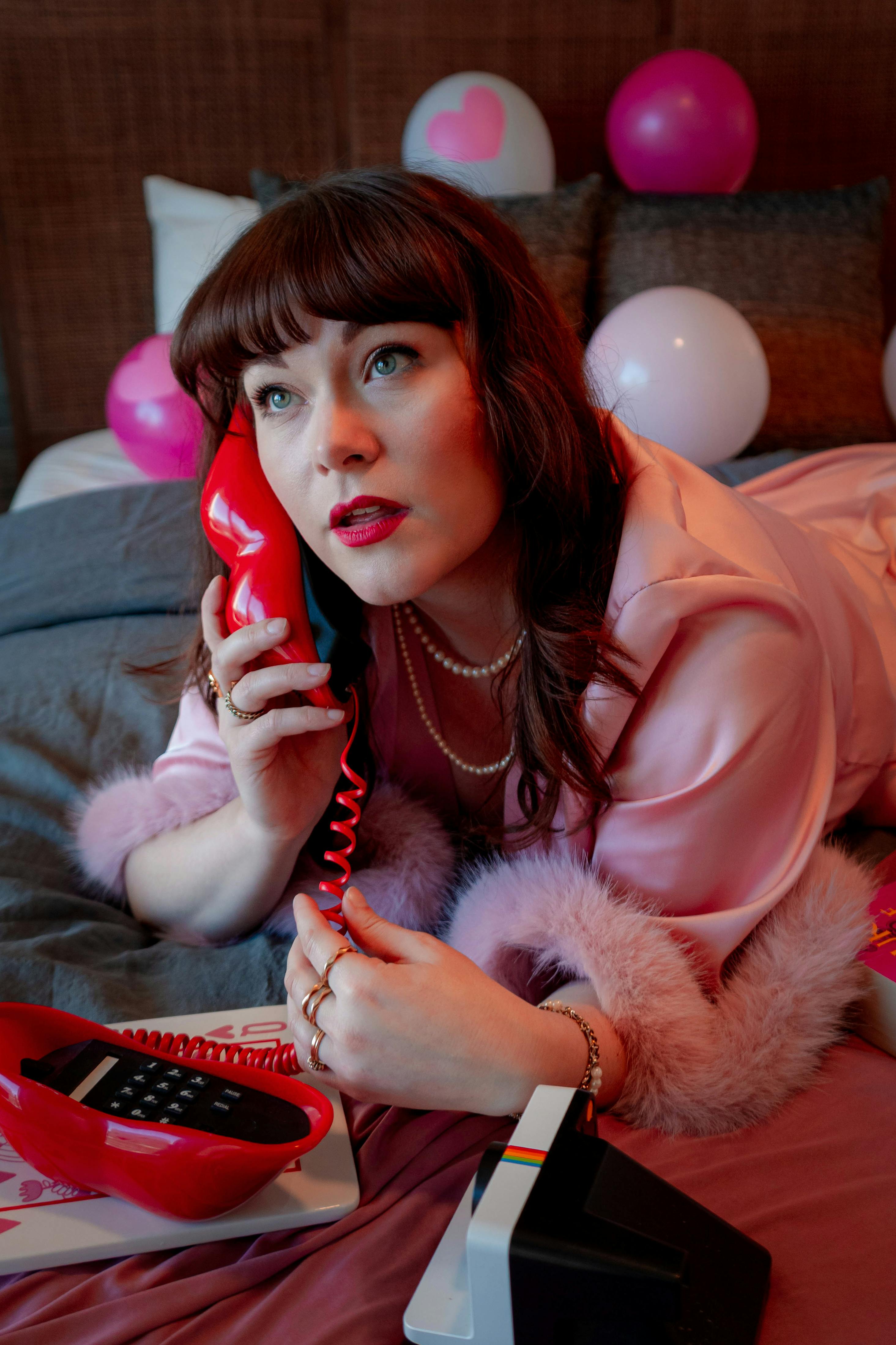 Free A woman in a pink outfit lies on a bed with a retro telephone and balloons. Stock Photo