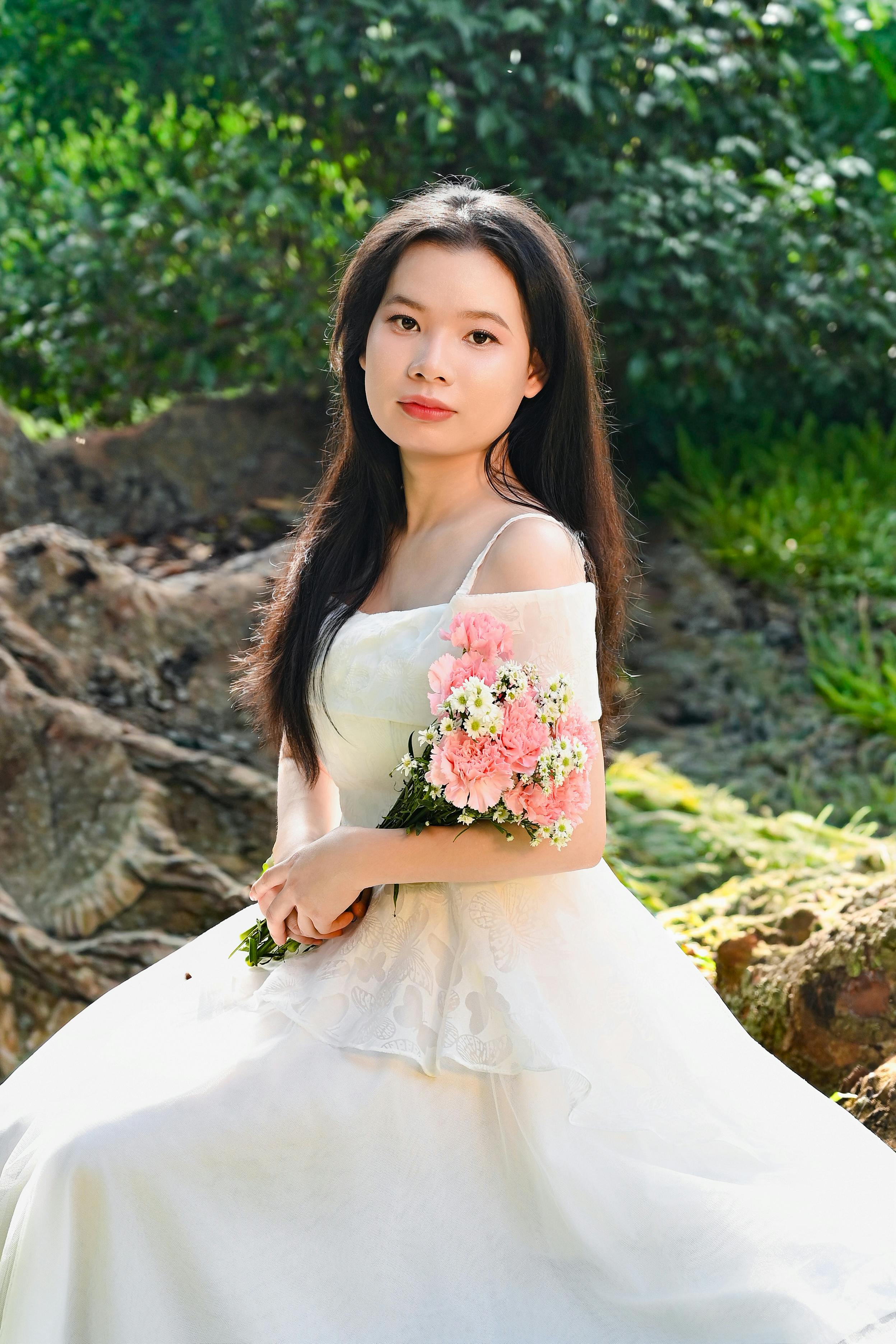 Gratis Elegante ritratto di una donna in abito bianco che tiene in mano dei fiori, seduta all'aperto in un giardino soleggiato. Foto a disposizione