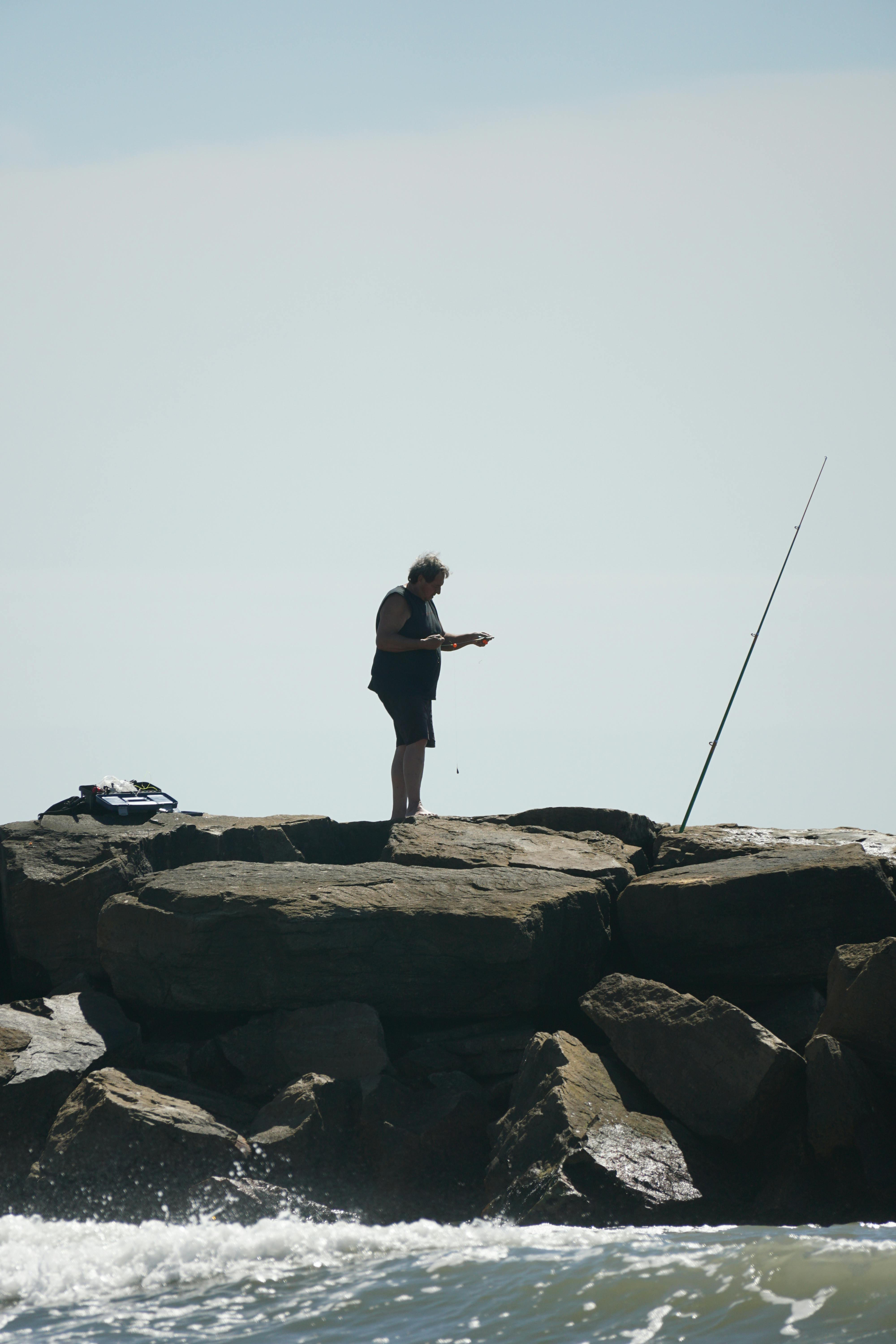 gratis Een eenzame man vist vanaf een rotsachtig strand onder een heldere hemel. Stockfoto