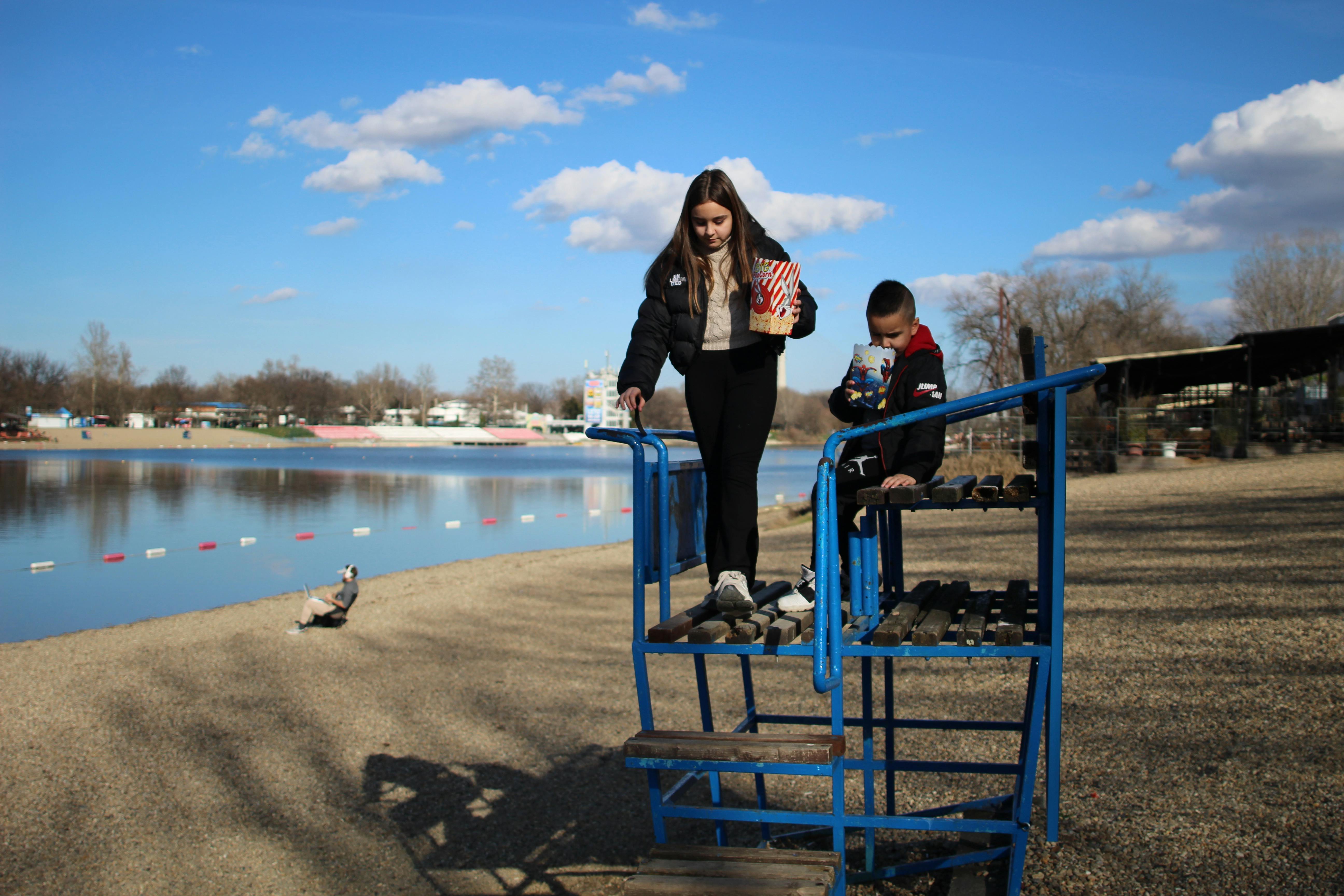 Δωρεάν Δύο παιδιά με ποπ κορν δίπλα σε μια λίμνη, απολαμβάνοντας μια ηλιόλουστη φθινοπωρινή μέρα. Φωτογραφία από στοκ φωτογραφιών