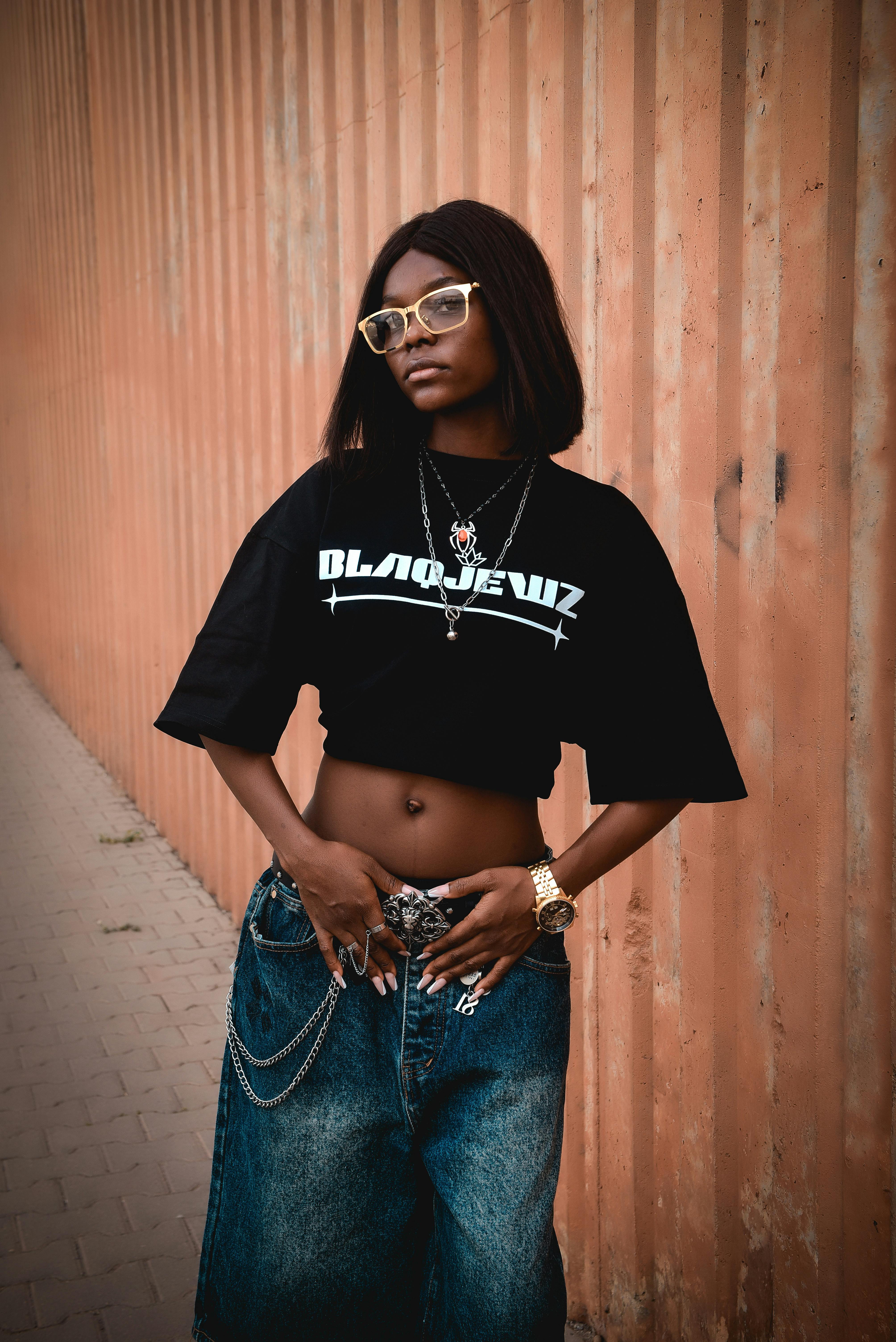 Gratis Giovane donna alla moda in elegante abbigliamento streetwear in posa contro un muro all'aperto. Foto a disposizione