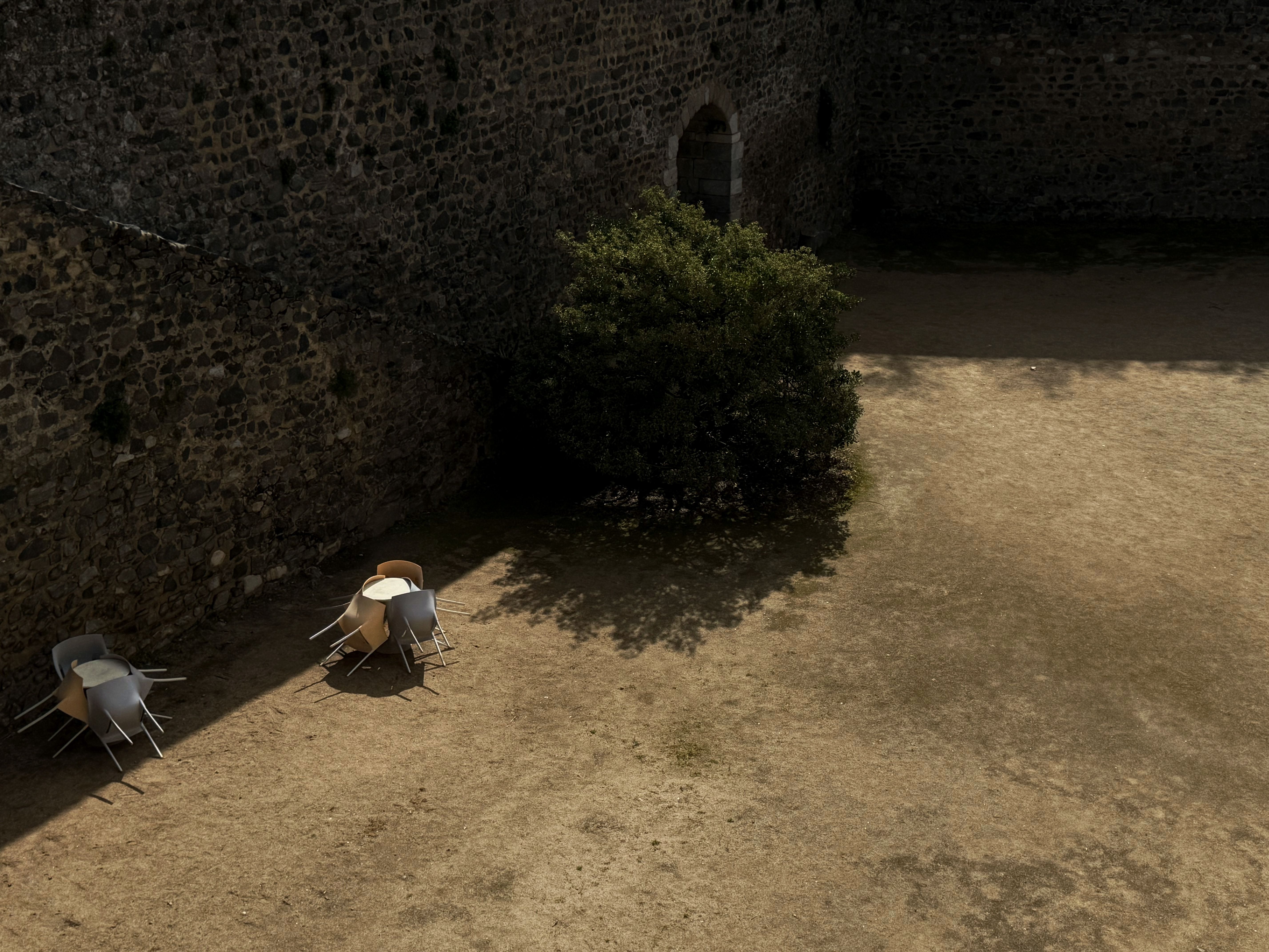 Empty courtyard with stone walls, tables, and chairs under natural sunlight casting long shadows.