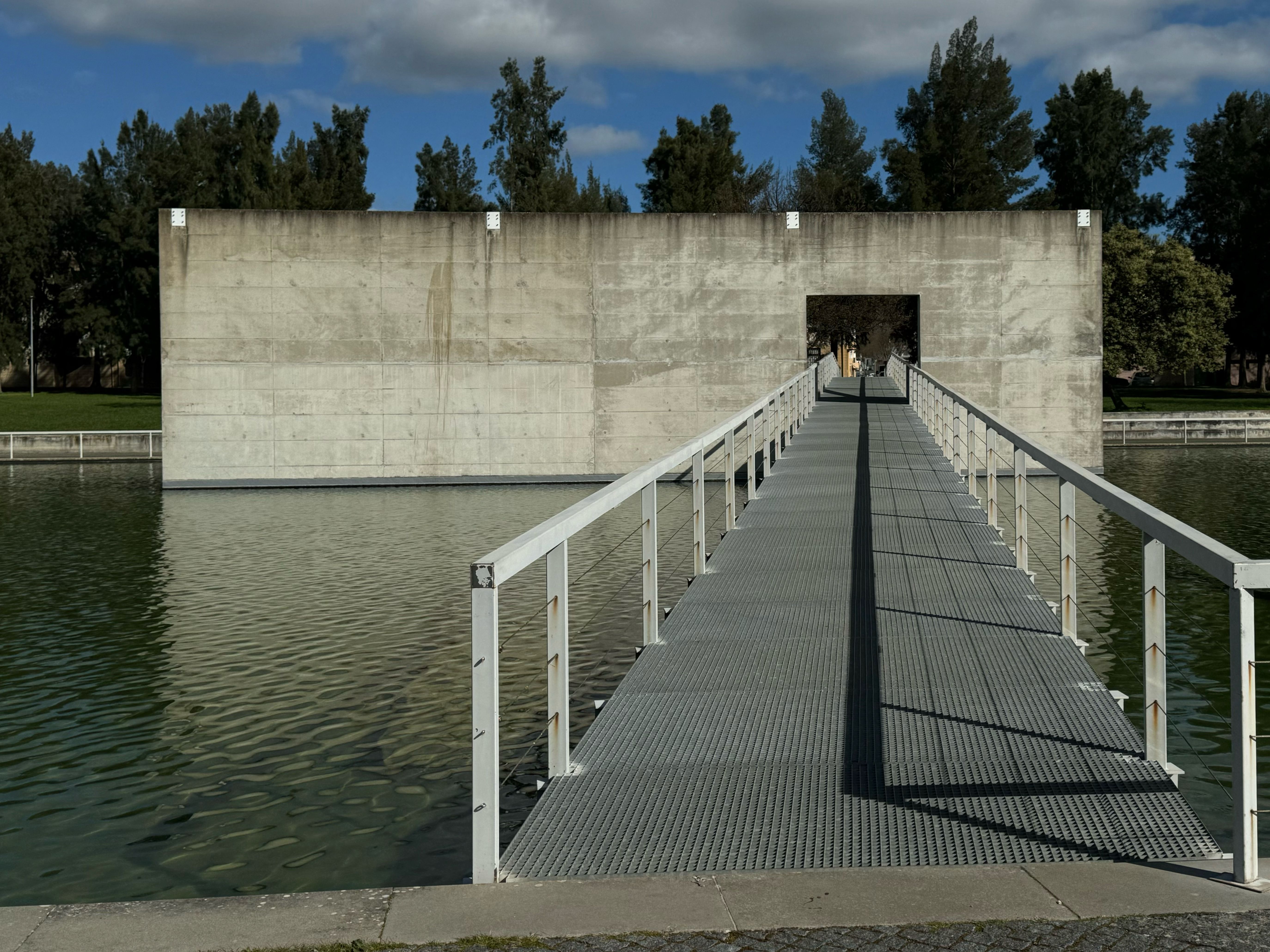 A modern bridge leads to a concrete structure over reflective water, surrounded by greenery under a bright sky.
