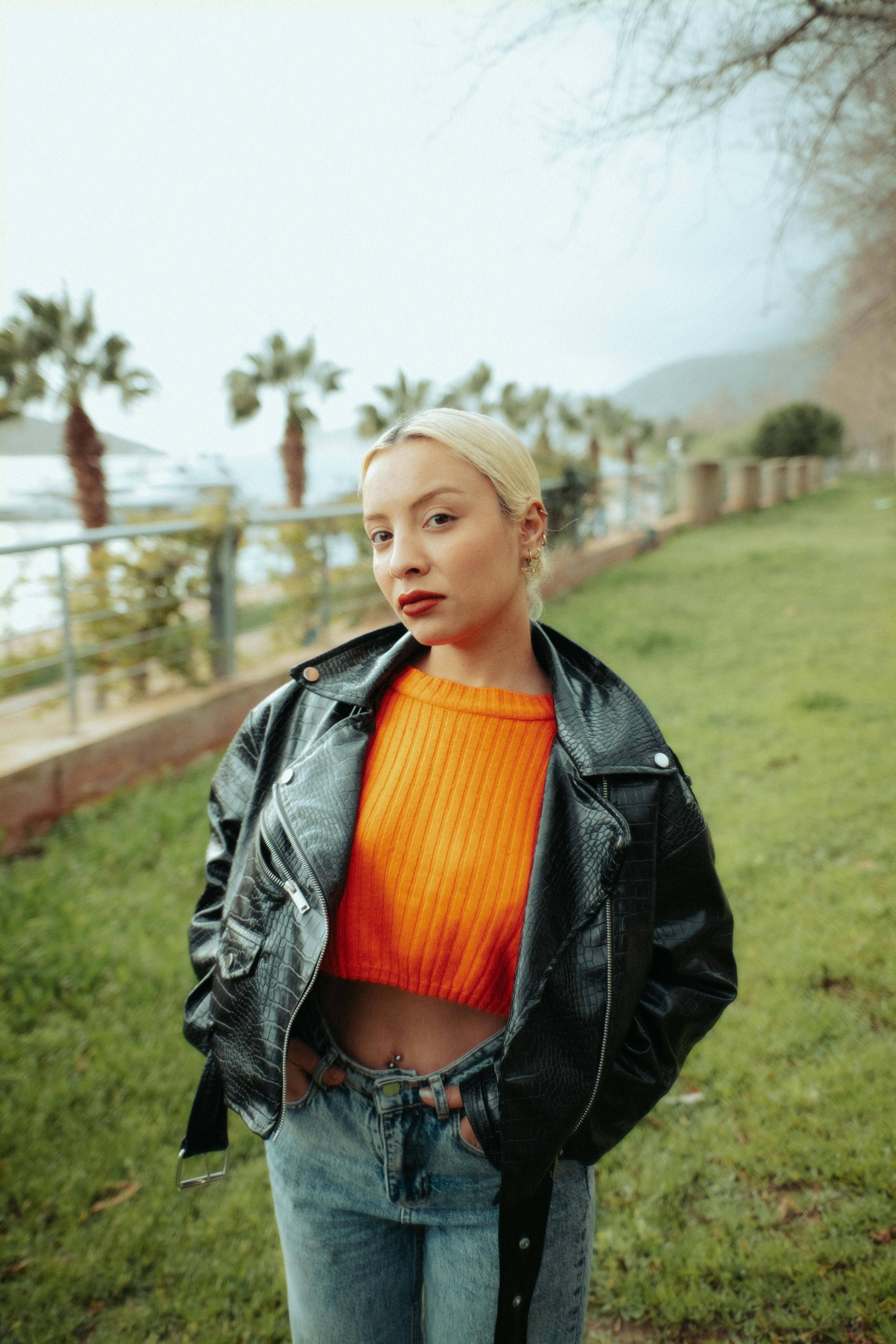 Δωρεάν Νέα γυναίκα που φορά ένα δερμάτινο μπουφάν και ένα πορτοκαλί πουλόβερ σε εξωτερικό χώρο δίπλα σε φοίνικες. Φωτογραφία από στοκ φωτογραφιών