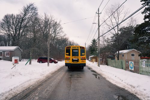 Free A school bus on a snowy rural road, winter landscape, trees, houses, and overcast sky. Stock Photo