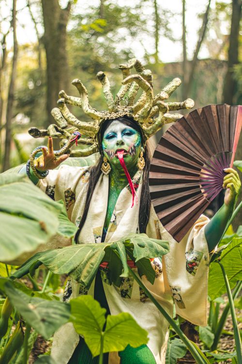 Free Colorful cosplay character with artistic makeup in a forest setting holding a fan. Stock Photo