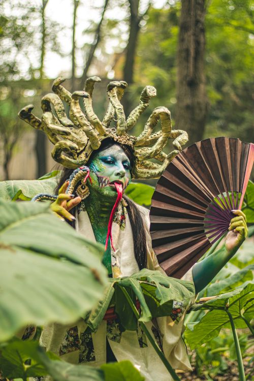 Free A colorful Gorgon cosplayer poses with fan amidst lush greenery, inspired by mythical and cultural themes. Stock Photo