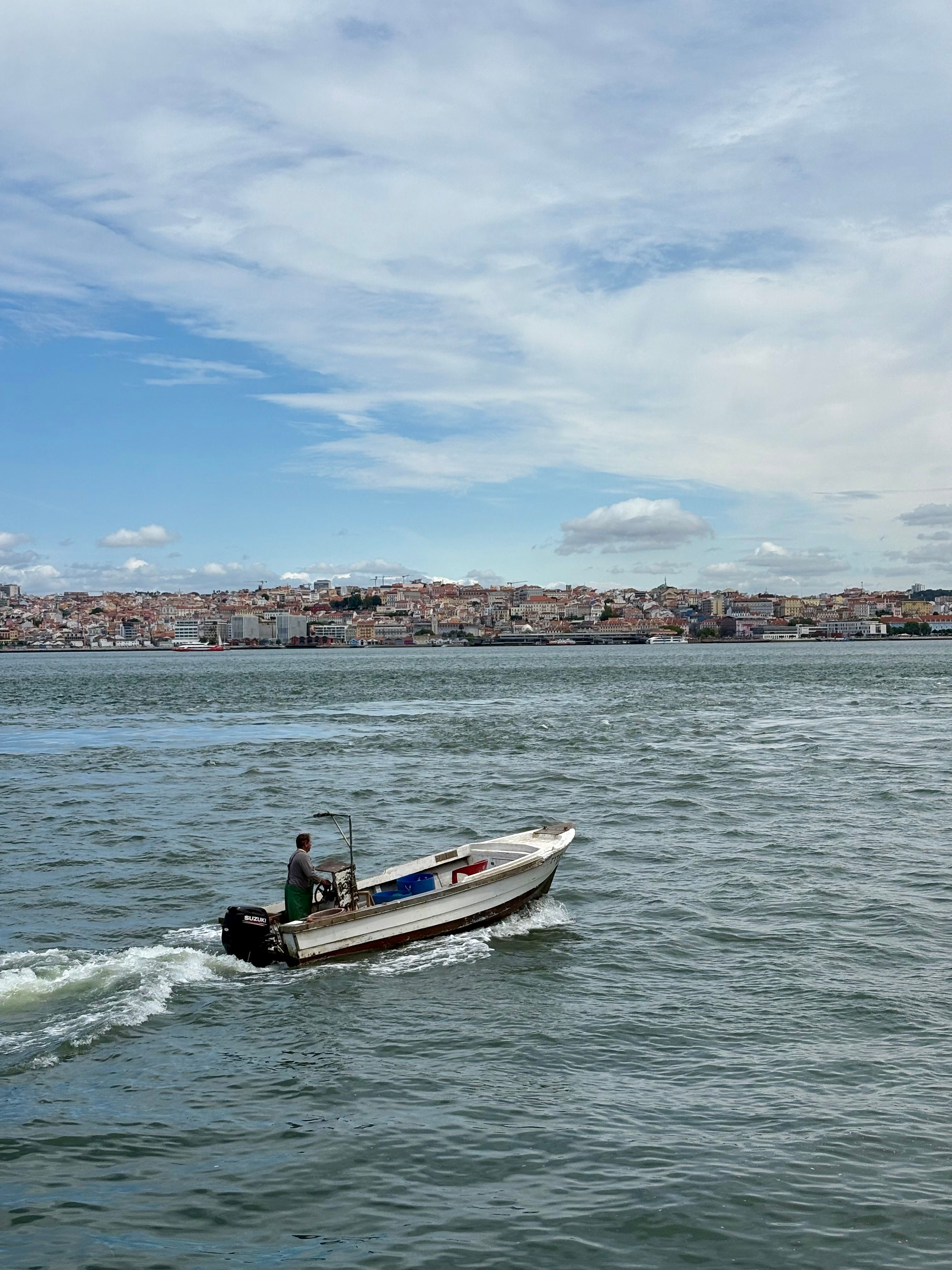A lone fisherman navigates his motorboat on the Tagus River with Almada's cityscape in the background.