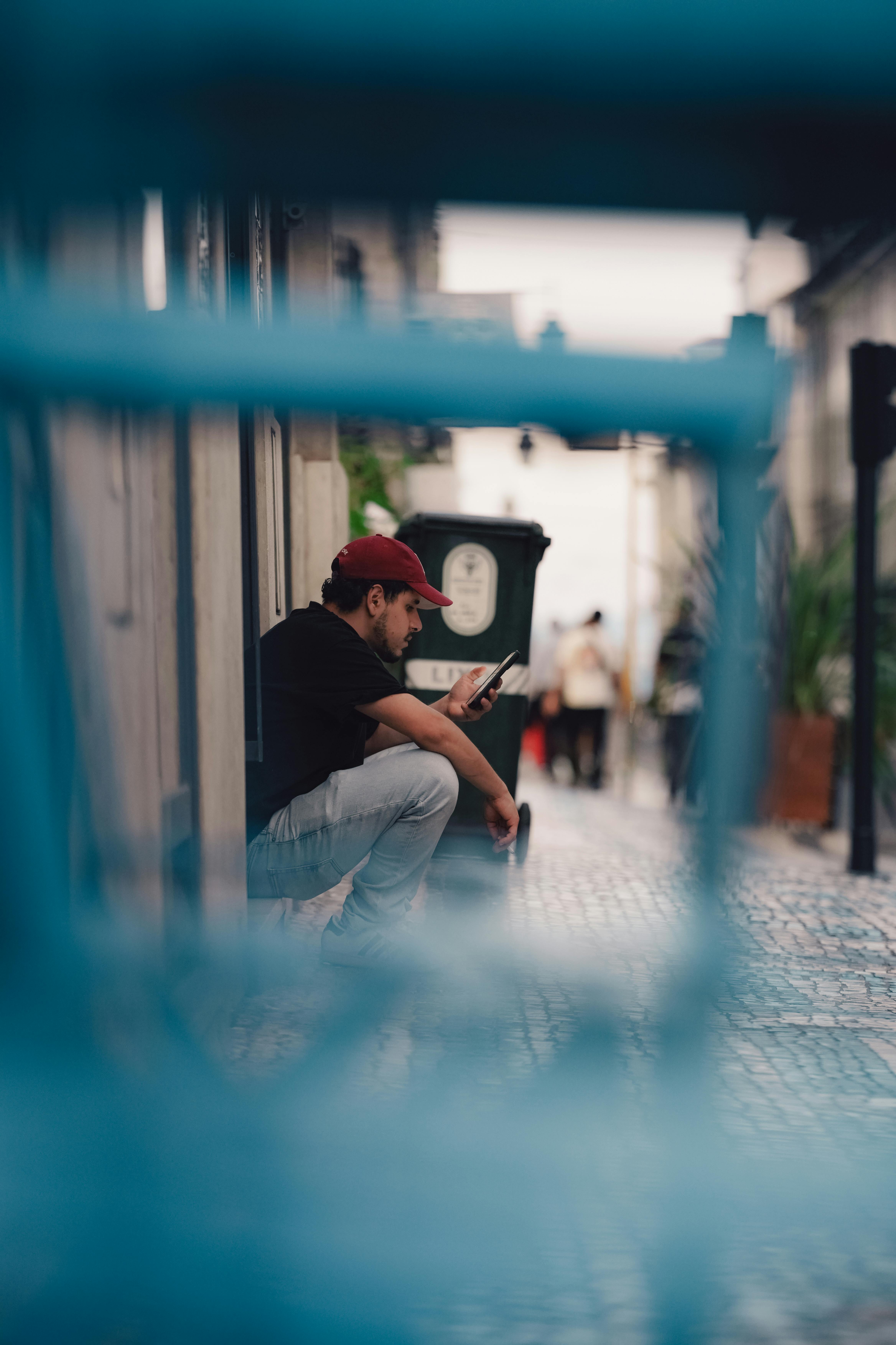 Gratis Un giovane uomo con un berretto rosso è seduto sul marciapiede e usa il telefono in un contesto urbano. Foto a disposizione