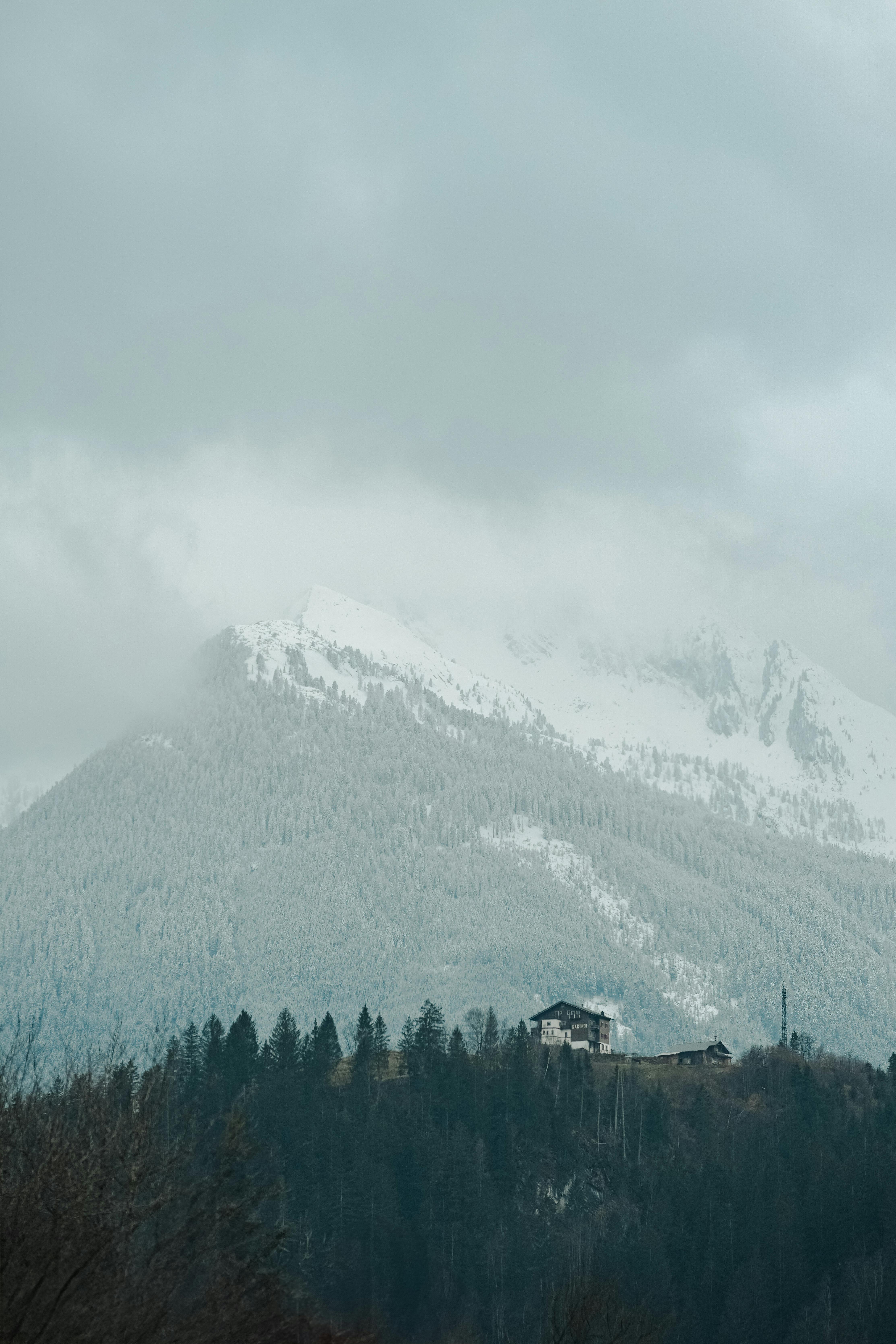 Δωρεάν Γραφική θέα ενός χιονισμένου βουνού με μια μακρινή καλύβα κάτω από συννεφιασμένους ουρανούς. Φωτογραφία από στοκ φωτογραφιών