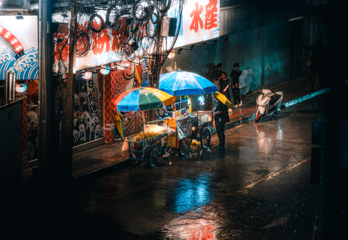 Colorful street food carts in rainy Bangkok night market scene.